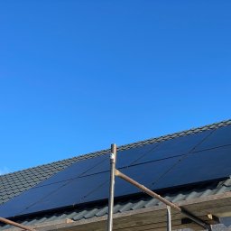 MAREK KALICIAK - Zielona Energia - Częściowo zamontowane czarne panele na szarym dachu z dachówki ceramicznej widziane spod rusztowania na tle błękitnego nieba.