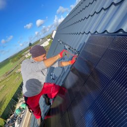 MAREK KALICIAK - Zielona Energia - Monter w czerwonych spodniach i rękawicach instaluje panel słoneczny na dachu z metalowej blachy w słoneczny dzień, widok na zielone pola w tle.