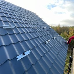 MAREK KALICIAK - Zielona Energia - Montażysta w czerwonym kombinezonie na dachu z szarej blachodachówki, instaluje aluminiowe profile pod panele fotowoltaiczne, widok z dołu na rusztowanie.