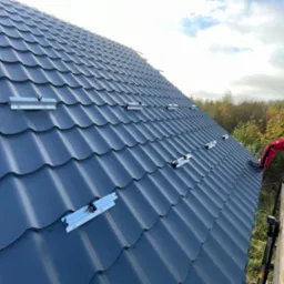 Montażysta w czerwonym kombinezonie na dachu z szarej blachodachówki, instaluje aluminiowe profile pod panele fotowoltaiczne, widok z dołu na rusztowanie.