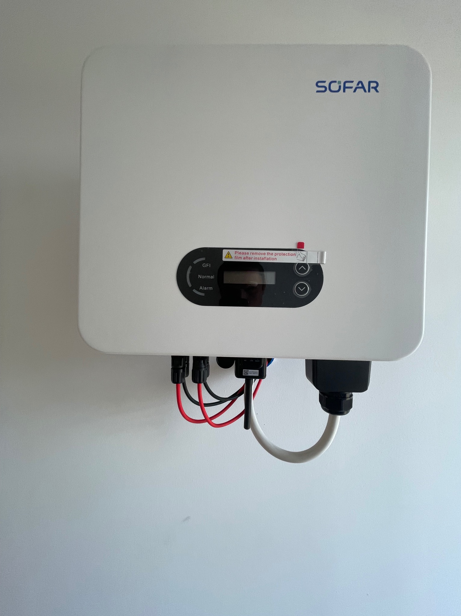 Biały falownik SOFAR zamontowany na ścianie, widoczne kable zasilające i naklejka z informacją o usunięciu folii ochronnej po instalacji.