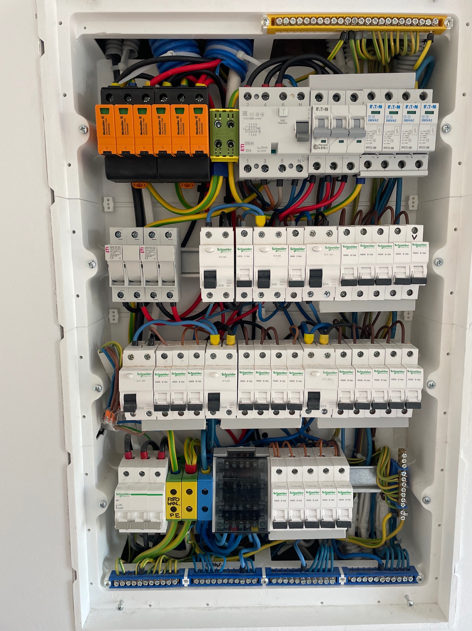 Rozdzielnica elektryczna z zabezpieczeniami przepięciowymi, bezpiecznikami, okablowaniem i oznaczeniami obwodów fotowoltaicznych, zamontowana w białej obudowie.