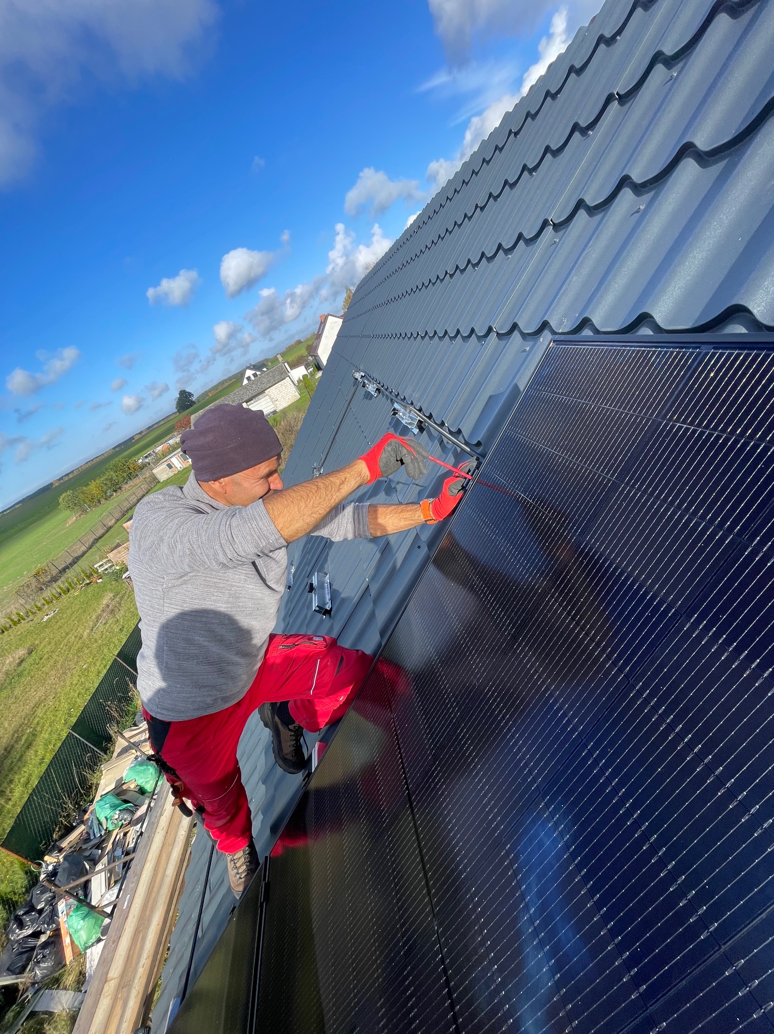 Monter w czerwonych spodniach i rękawicach instaluje panel słoneczny na dachu z metalowej blachy w słoneczny dzień, widok na zielone pola w tle.