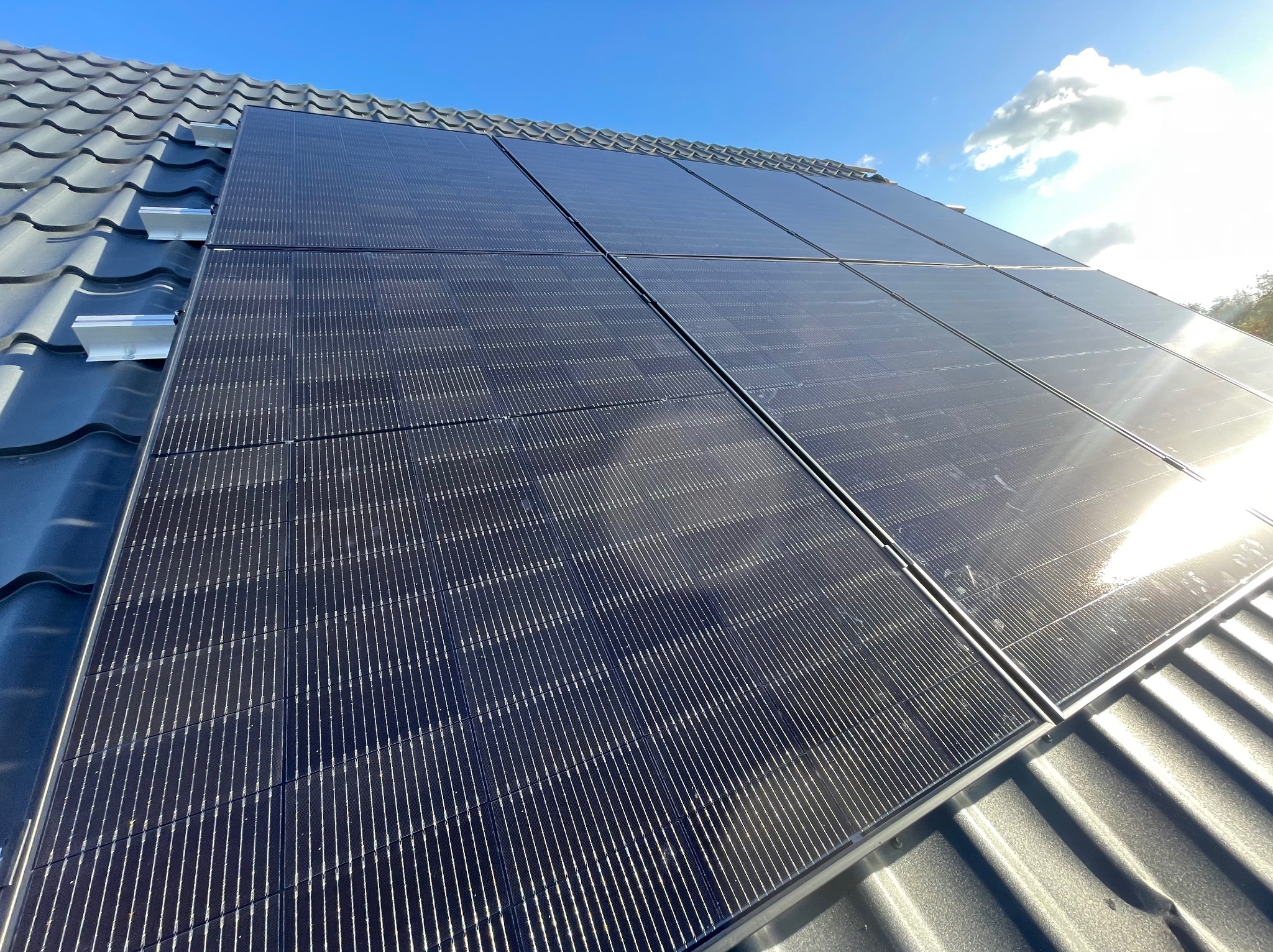 Panele słoneczne zamontowane na szarym dachu z dachówki ceramicznej, widoczne mocowania paneli, błękitne niebo w tle.