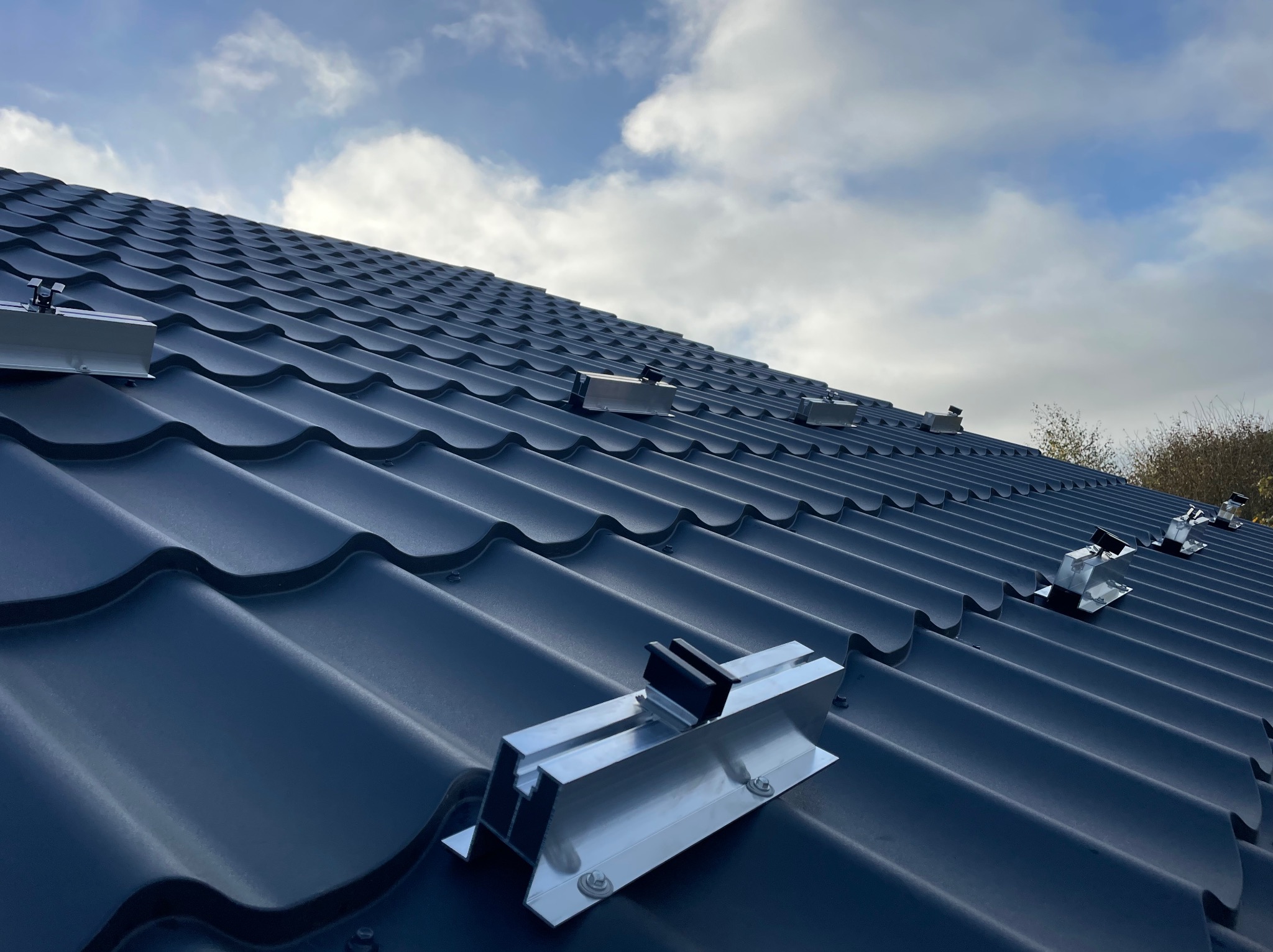 Metalowe uchwyty montażowe na ciemnoszarej dachówce ceramicznej w przygotowaniu do instalacji paneli słonecznych, widoczne śruby mocujące, pochmurne niebo w tle.
