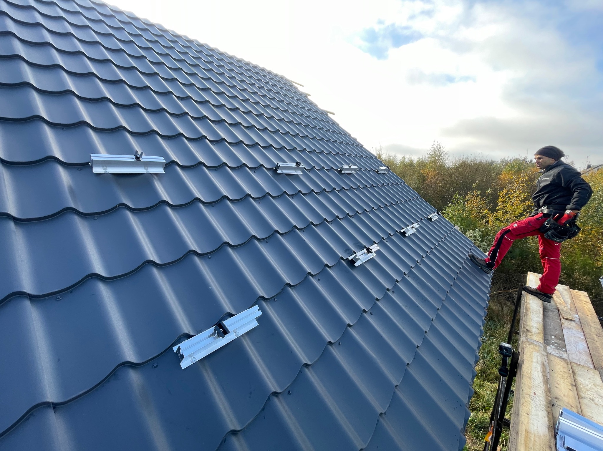 Montażysta w czerwonym kombinezonie na dachu z szarej blachodachówki, instaluje aluminiowe profile pod panele fotowoltaiczne, widok z dołu na rusztowanie.