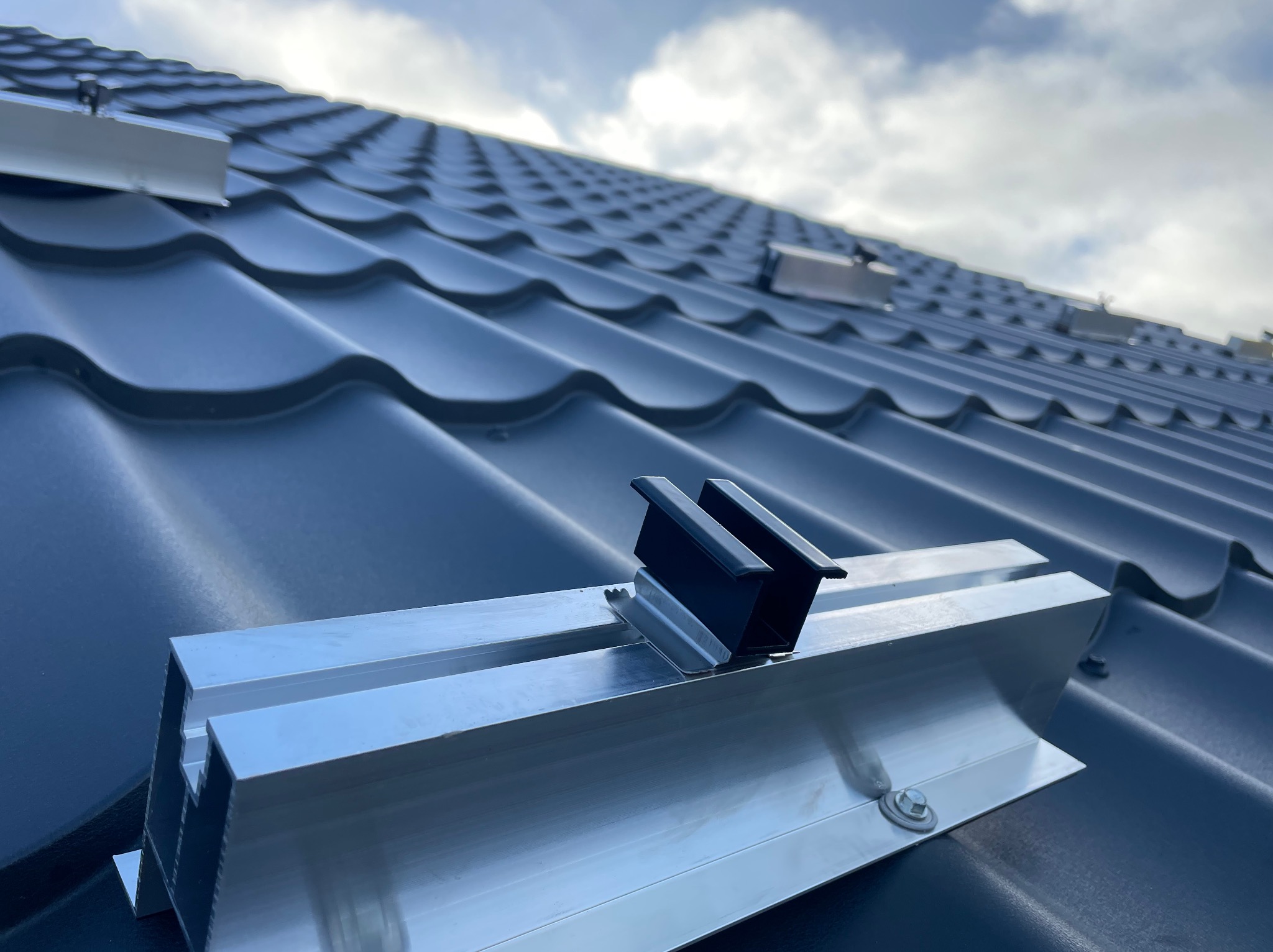 Szyny montażowe z uchwytami na panel słoneczny, zamocowane do grafitowej blachodachówki na tle pochmurnego nieba.