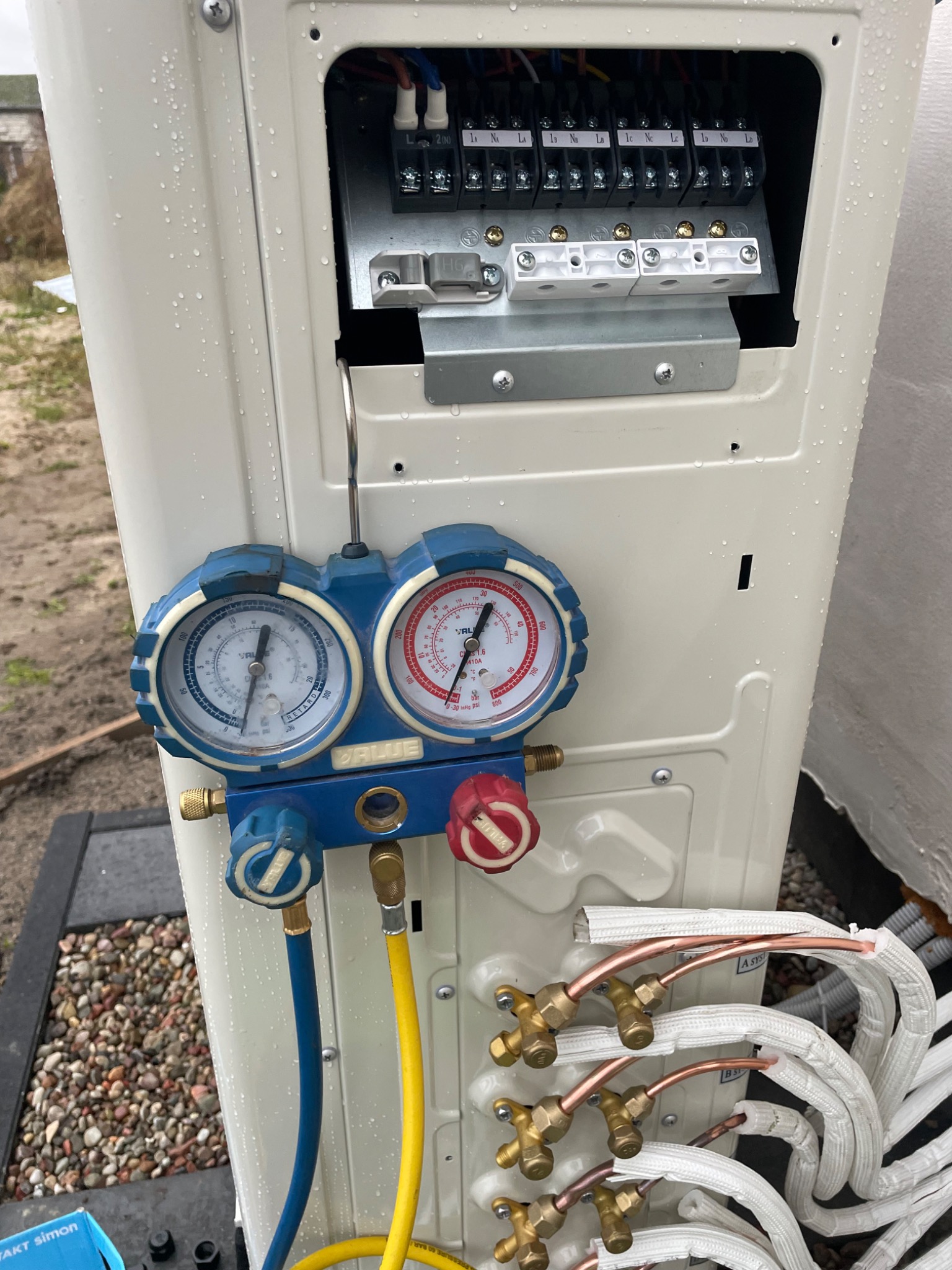 Podłączenie manometrów serwisowych do zewnętrznej jednostki klimatyzacji podczas montażu, widoczne kable elektryczne i rury chłodnicze w izolacji.