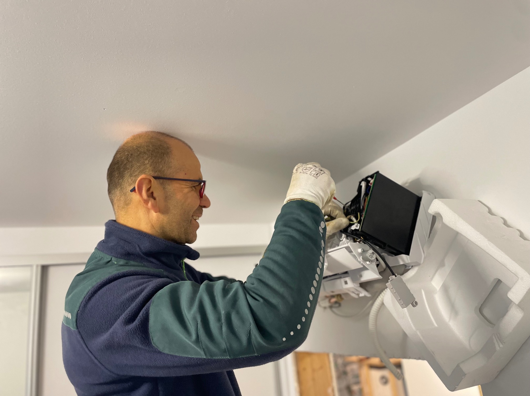 Uśmiechnięty instalator w okularach i rękawicach roboczych montuje jednostkę wewnętrzną klimatyzacji na białej ścianie, widoczne przewody i elementy montażowe.