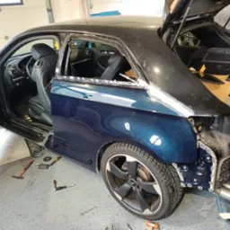 Niebieski Audi A3 bez tylnej szyby i fragmentu karoserii, widoczne narzędzia i elementy blacharskie w warsztacie.