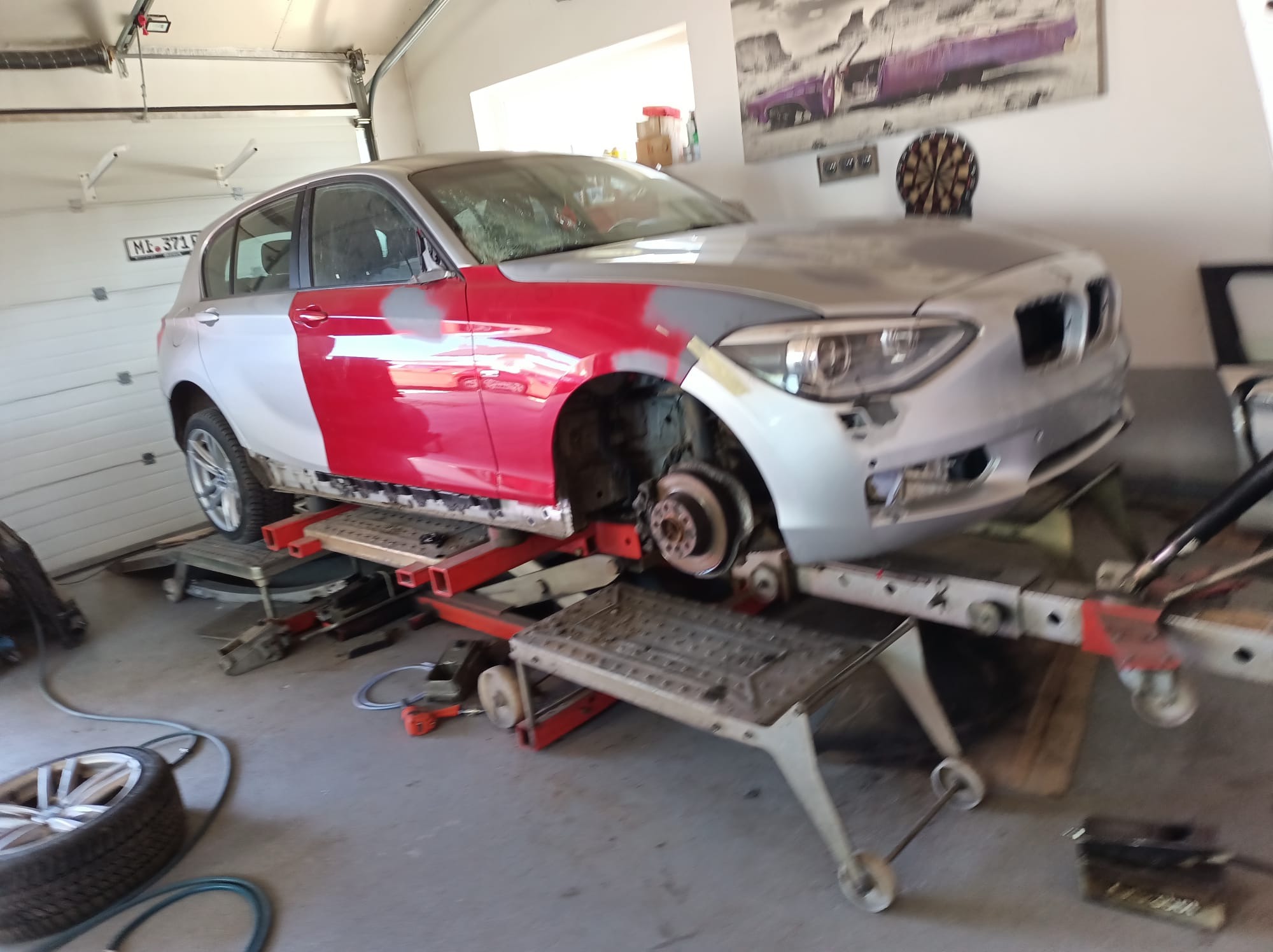 Samochód BMW na podnośniku w garażu, z fragmentami karoserii w czerwonym podkładzie i szarym szpachlu, przygotowywany do lakierowania.