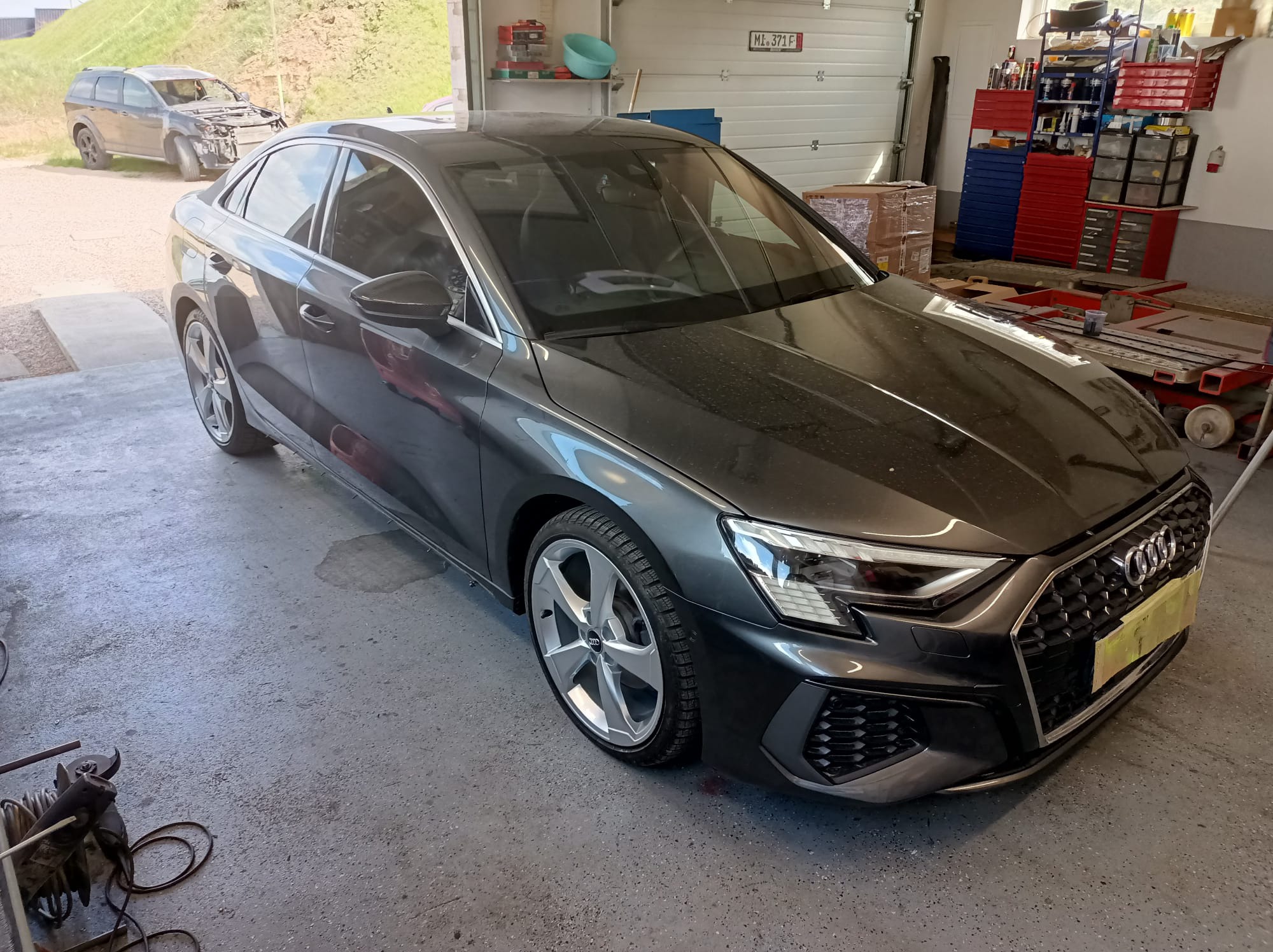 Szary samochód Audi A3 po naprawie blacharskiej, stojący w warsztacie z podnośnikiem i narzędziami w tle, z innym uszkodzonym pojazdem widocznym na zewnątrz.