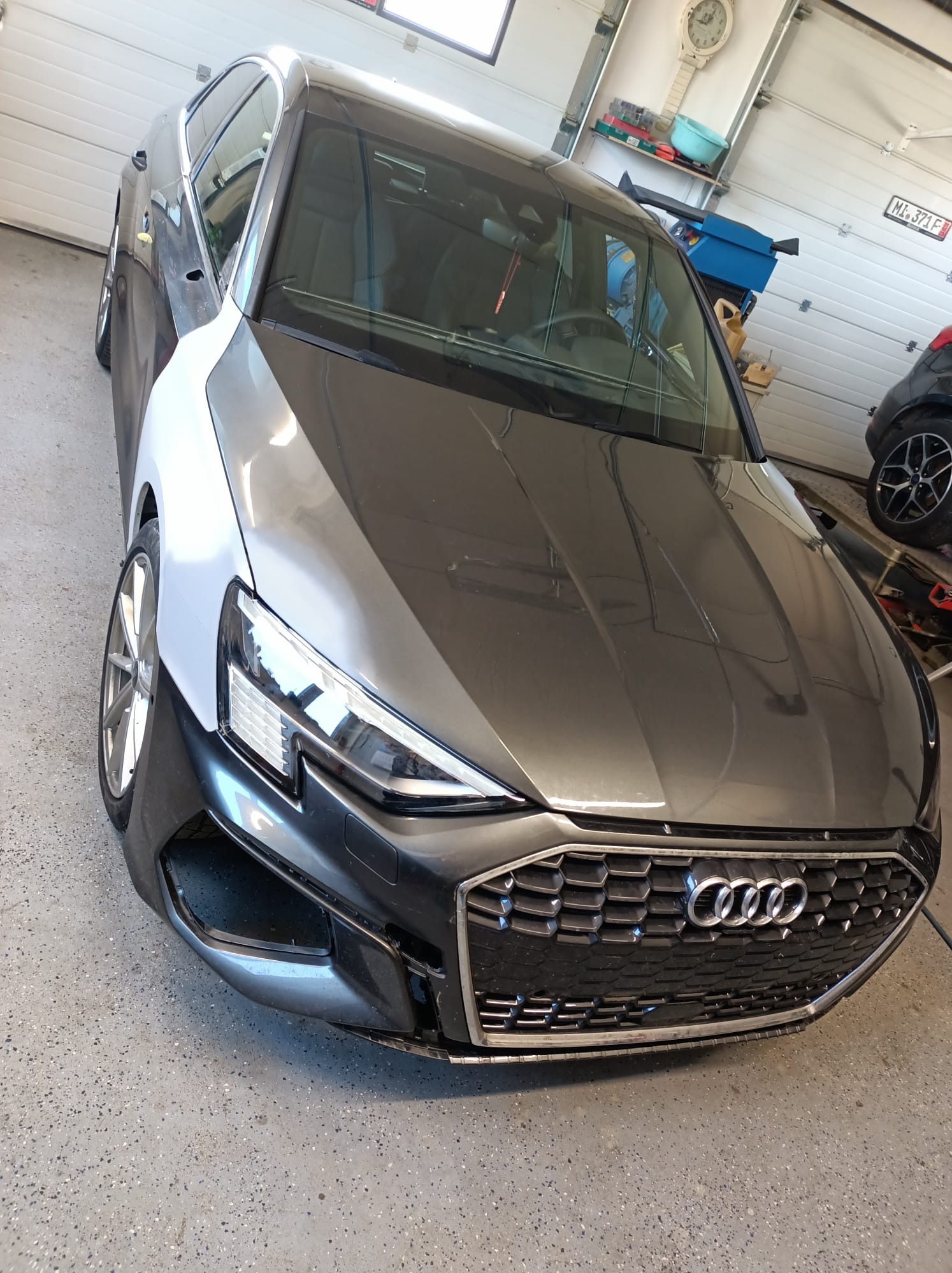 Szary samochód Audi z elementami karoserii w jasnym podkładzie, widoczny w warsztacie z zegarem na ścianie.