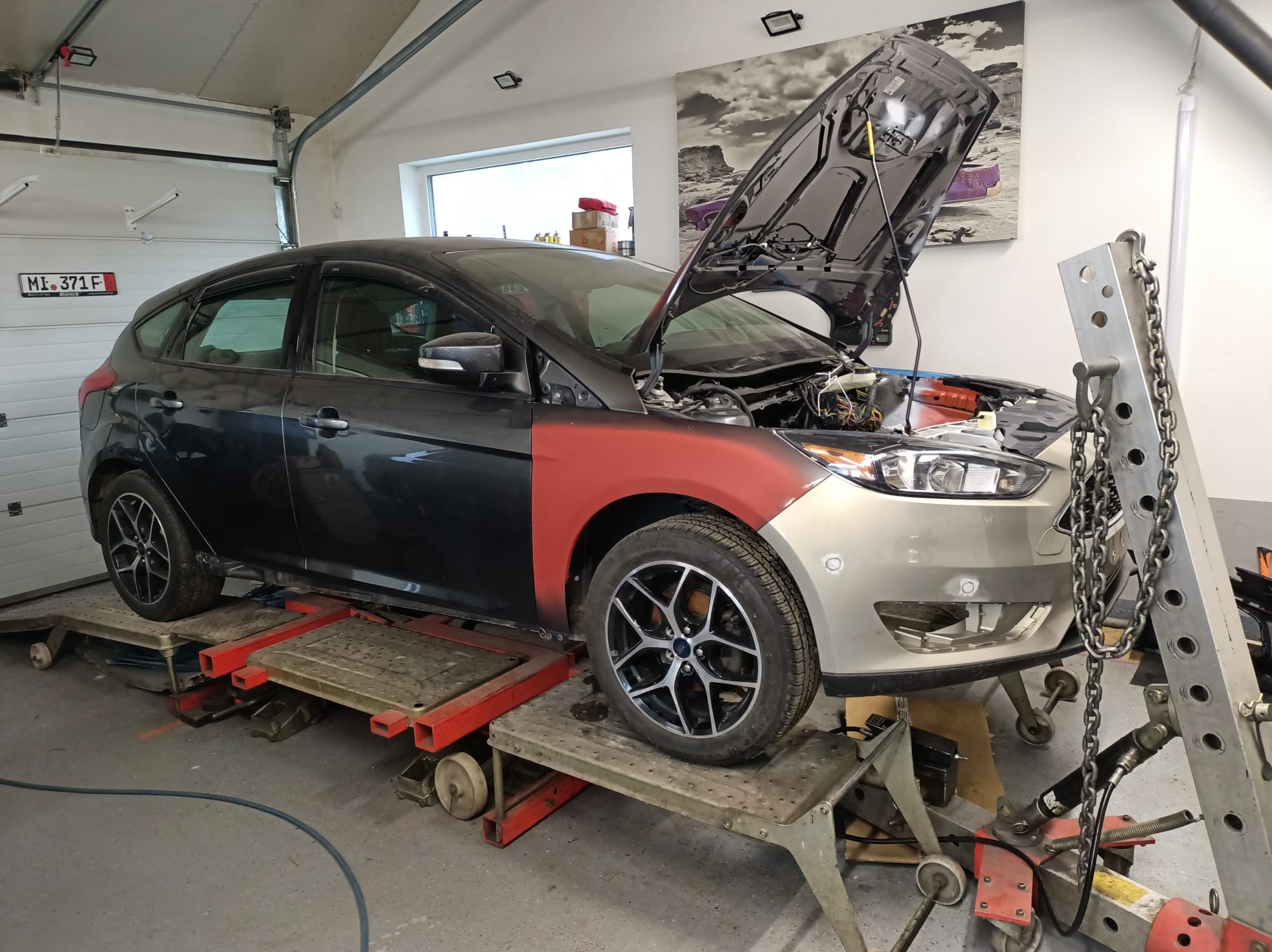 Samochód na platformie warsztatowej z otwartą maską, w trakcie naprawy karoserii, z widocznym fragmentem pomalowanym na czerwono i narzędziami blacharskimi obok.