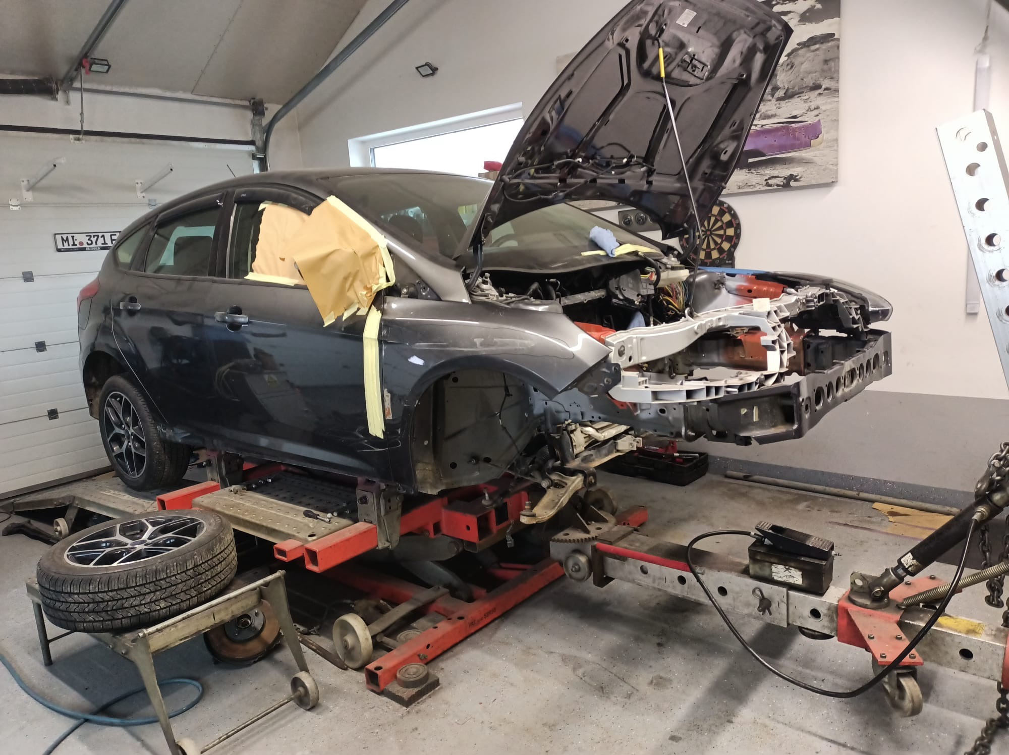 Szary samochód na czerwonym podnośniku w warsztacie, z otwartą maską i zdemontowanym przodem, zabezpieczony taśmą maskującą, widoczne narzędzia i koło na stojaku.