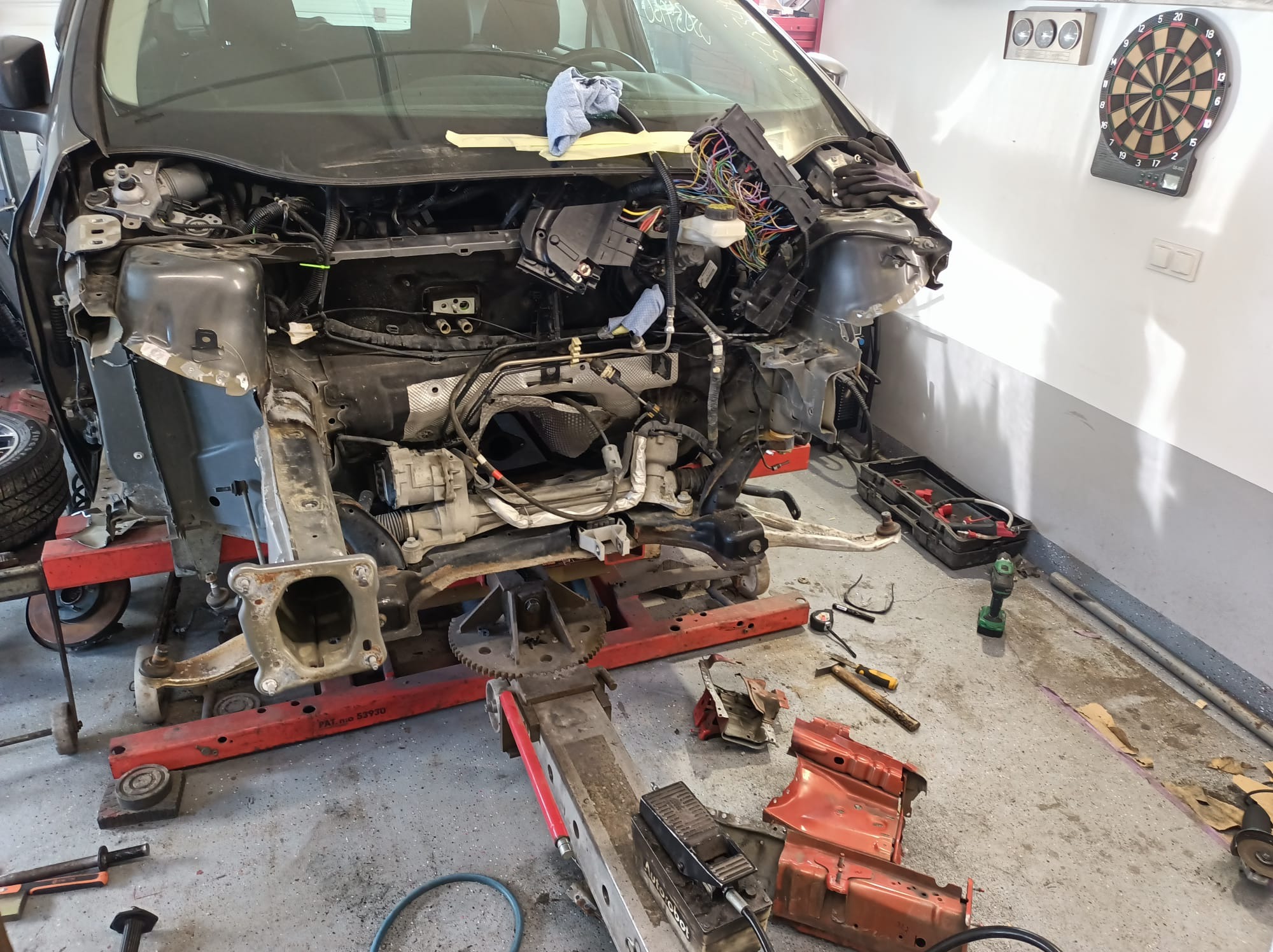 Uszkodzony samochód na stanowisku naprawczym z widocznymi elementami karoserii, przewodami elektrycznymi i narzędziami, w tle tarcza do darta.