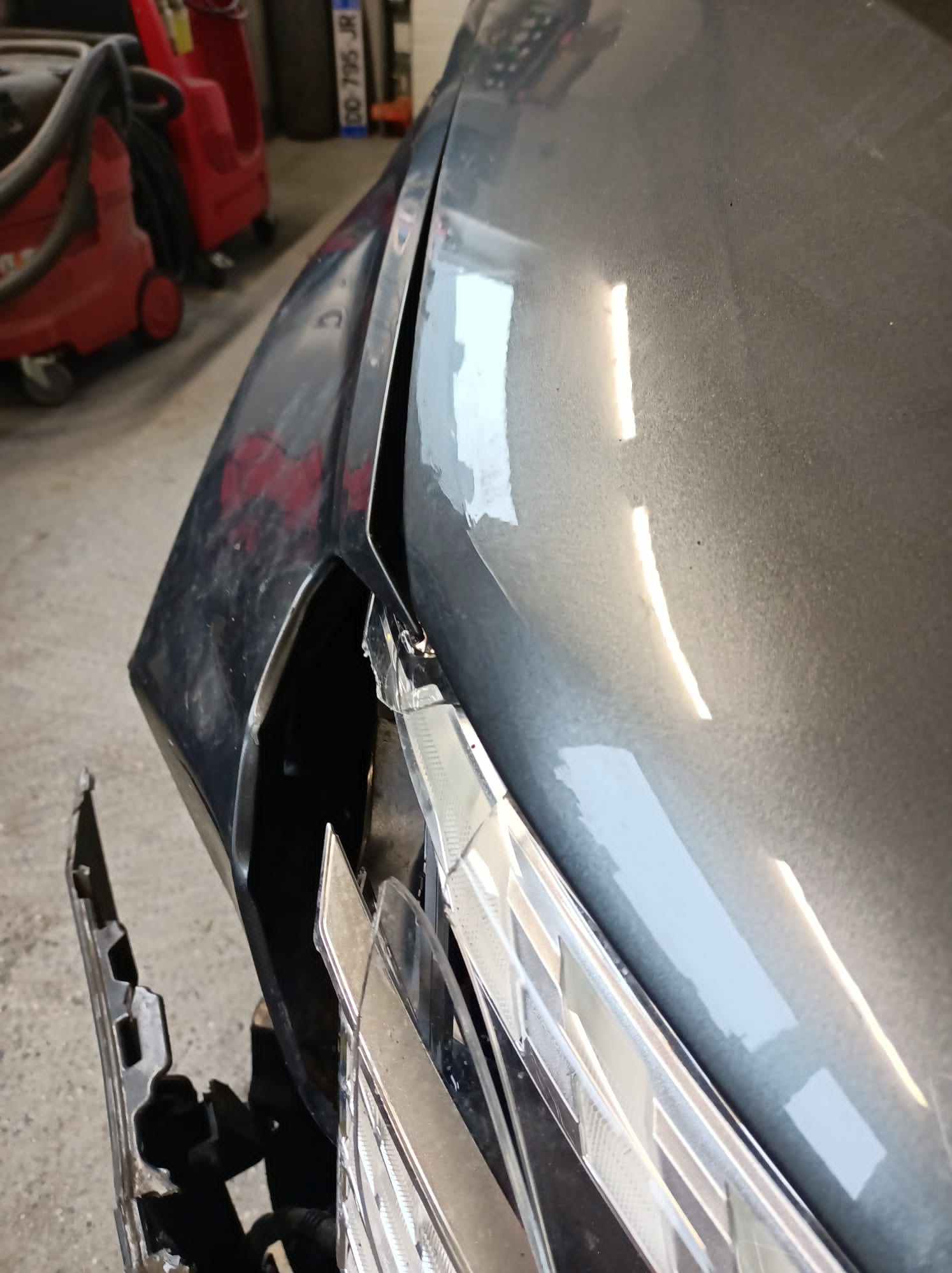 Uszkodzony, szary przedni zderzak samochodu z widocznymi pęknięciami i odwarstwiającym się lakierem, fragment uszkodzonej lampy widoczny w dolnej części kadru, w tle czerwony odkurzacz przemysłowy.