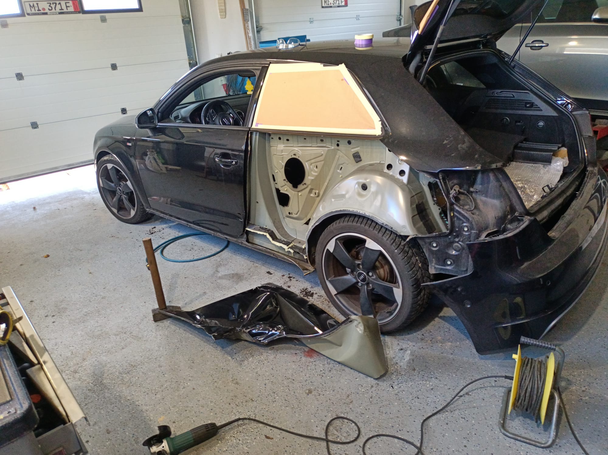 Czarny samochód Audi A3 w trakcie naprawy blacharskiej; brakujące elementy karoserii, widoczne zabezpieczenie okna taśmą, szlifierka kątowa i odłamki blachy na podłodze garażu.