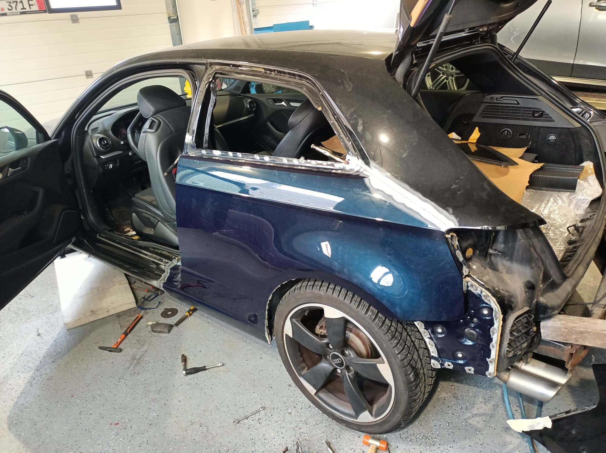 Niebieski Audi A3 bez tylnej szyby i fragmentu karoserii, widoczne narzędzia i elementy blacharskie w warsztacie.