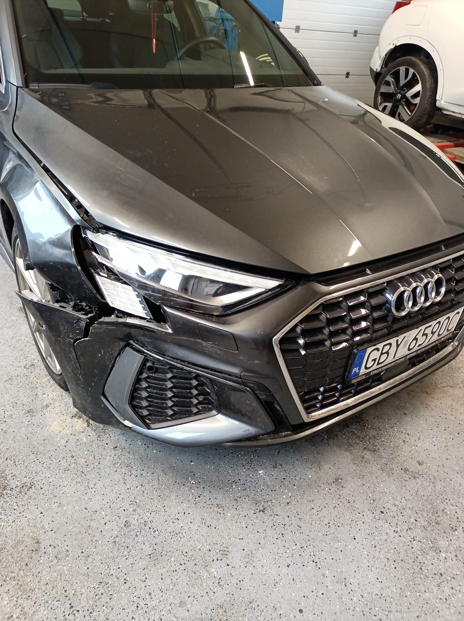 Uszkodzony przód szarego samochodu Audi z widocznym pękniętym zderzakiem, zarysowanym błotnikiem i uszkodzonym reflektorem, na tle białej bramy garażowej i częściowo widocznego innego pojazdu.