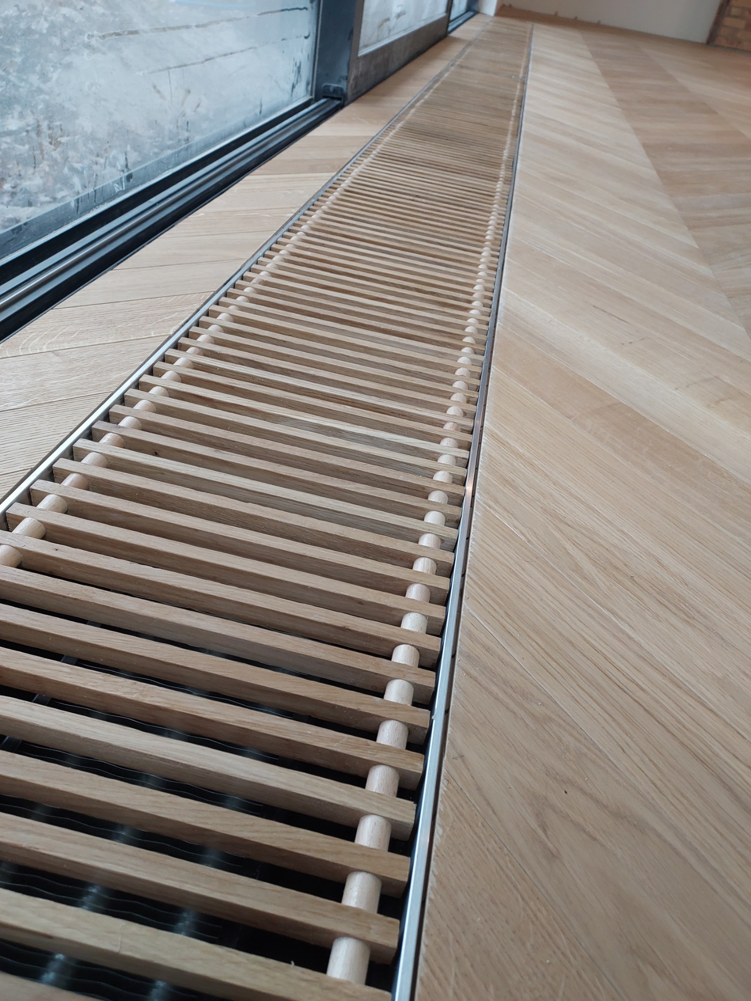 Jasny parkiet ułożony we wzór jodełki francuskiej, z wbudowanym w podłogę, drewnianym, liniowym grillem wentylacyjnym przy oknie.