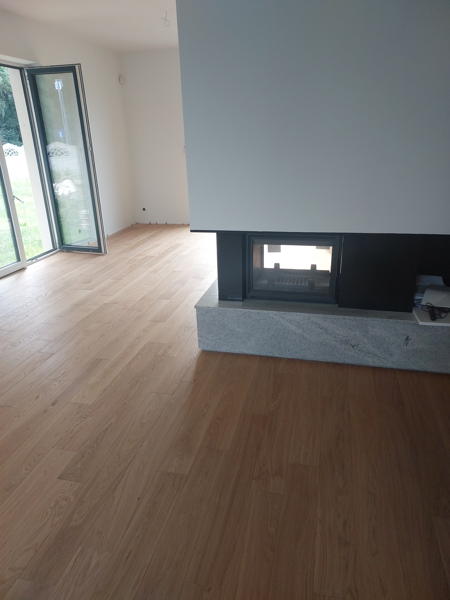 Jasny parkiet ułożony w nowoczesnym salonie z kominkiem, widoczne duże okno przesuwne.