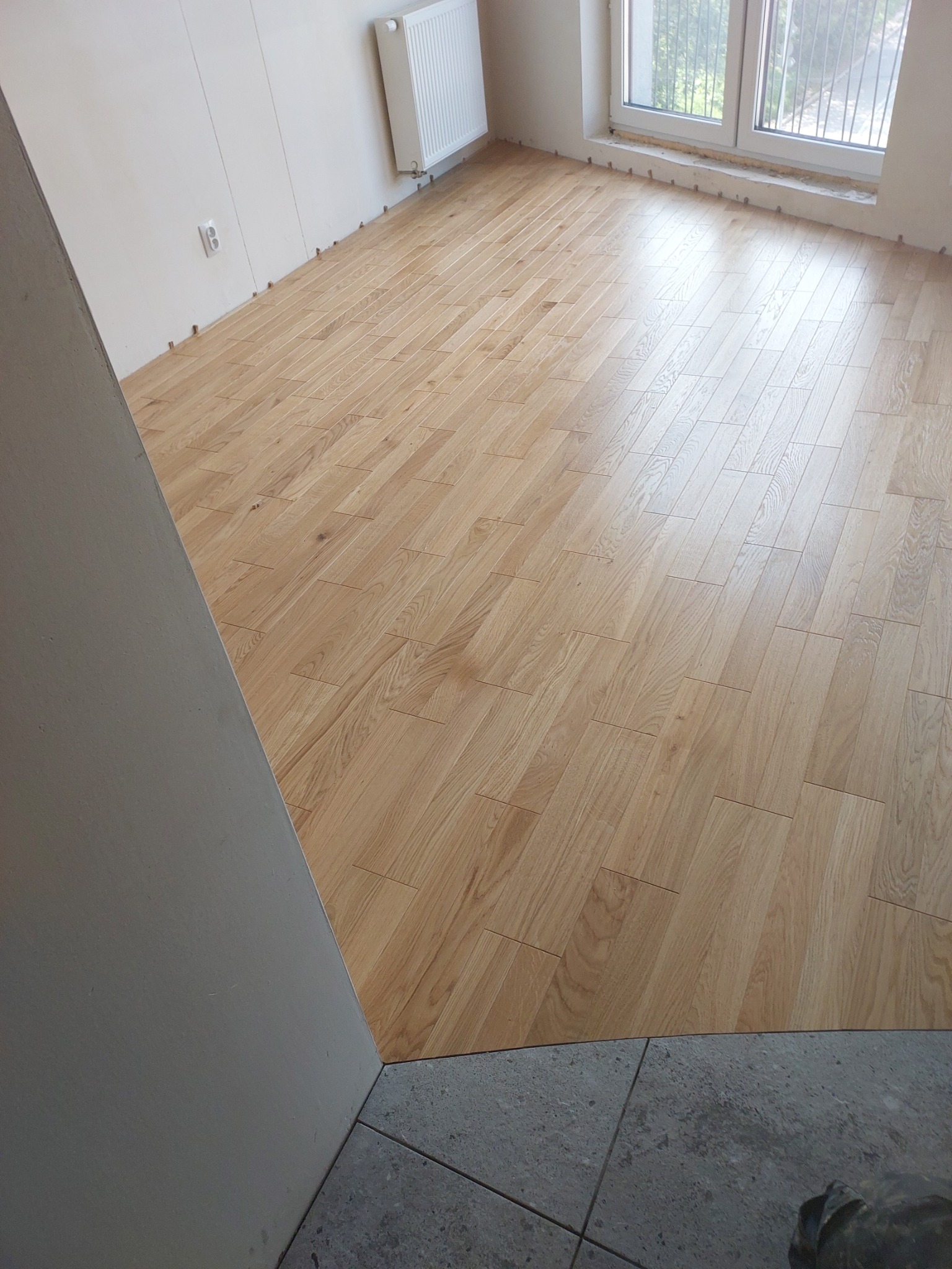 Jasne panele podłogowe ułożone w nowym pomieszczeniu, widoczne listwy przypodłogowe i grzejnik na ścianie.