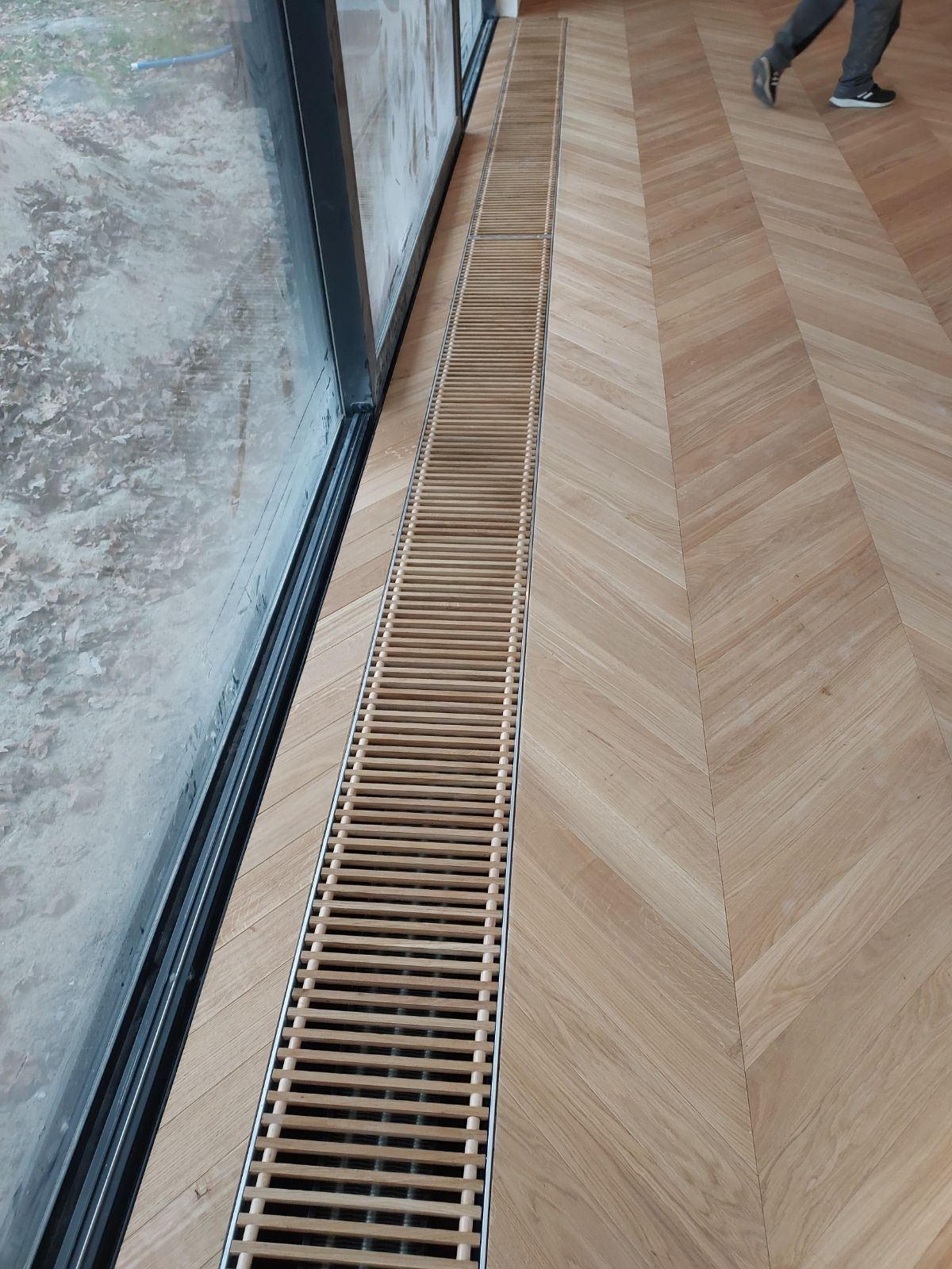 Jasny parkiet ułożony we wzór jodełki francuskiej, z wbudowanym w podłogę, liniowym, drewnianym kanałem grzewczym przy oknie.