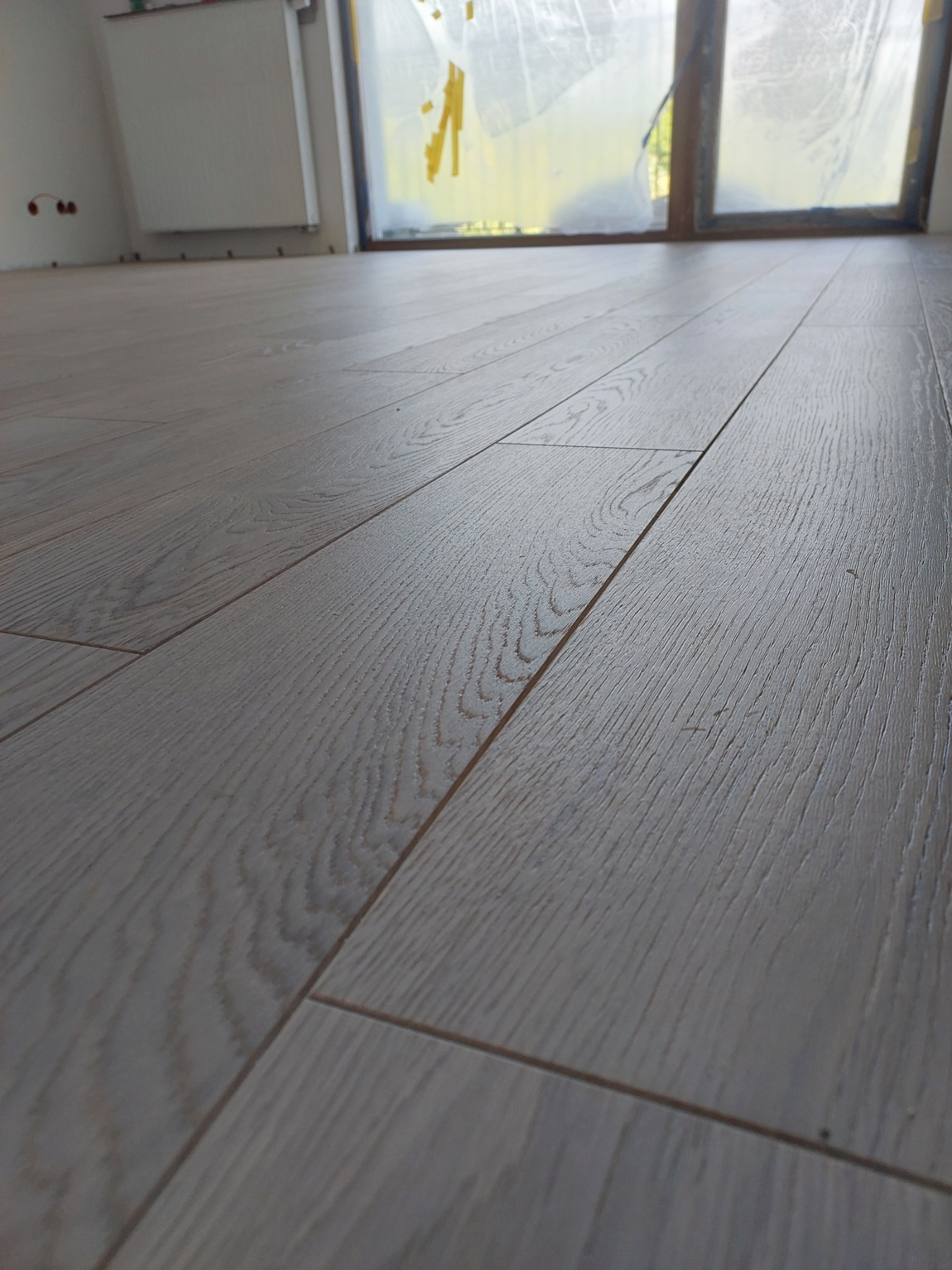 Jasne, drewniane panele podłogowe ułożone w nowym wnętrzu, widoczne okno z folią ochronną i fragment grzejnika.