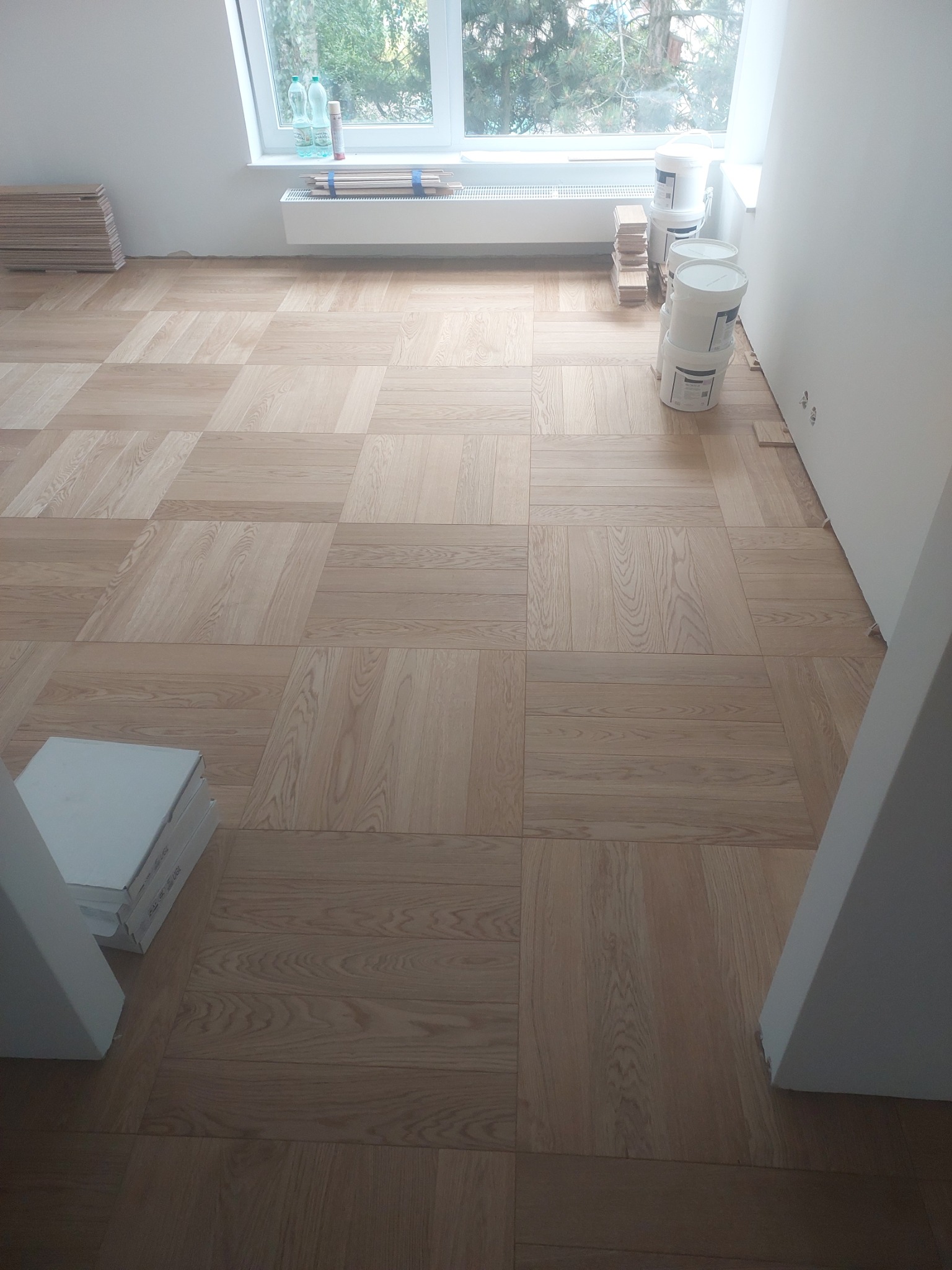 Wnętrze pokoju z ułożonym parkietem w kwadraty, widoczne puszki z farbą, paczki paneli i okno z widokiem na zieleń.