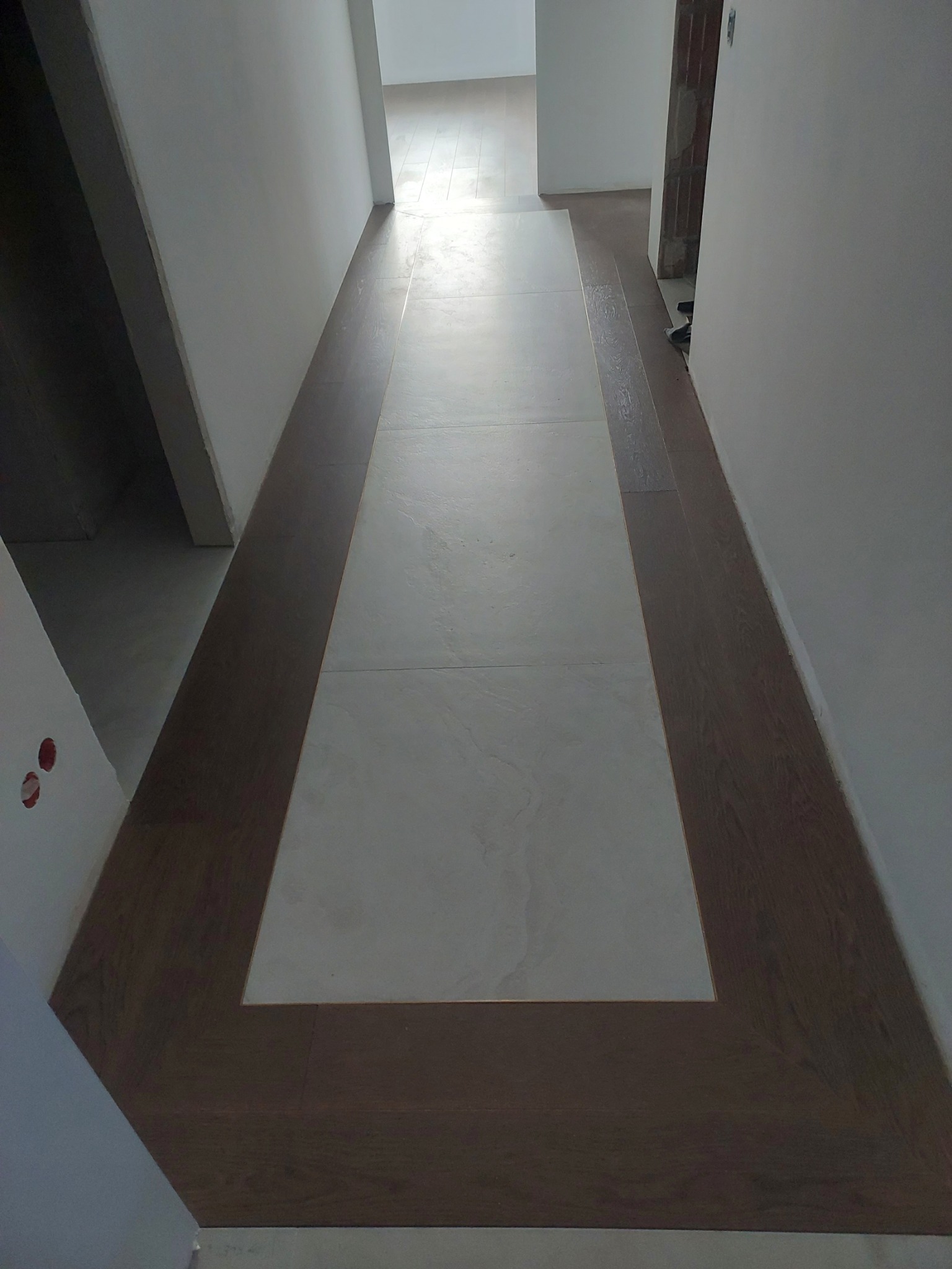 Korytarz z wykończoną podłogą: białe płytki otoczone ciemnymi panelami imitującymi drewno, widok z góry, surowy stan ścian.