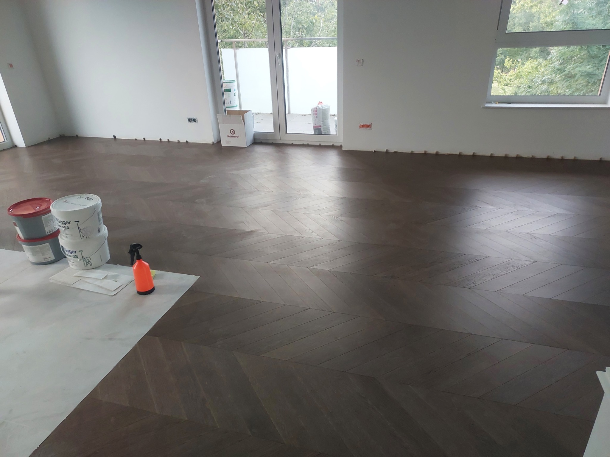 Ukończona podłoga z ciemnego parkietu ułożonego we wzór jodełki francuskiej, widoczne puszki z lakierem i spryskiwacz na zabezpieczonej powierzchni, listwy przypodłogowe jeszcze nie zamontowane.