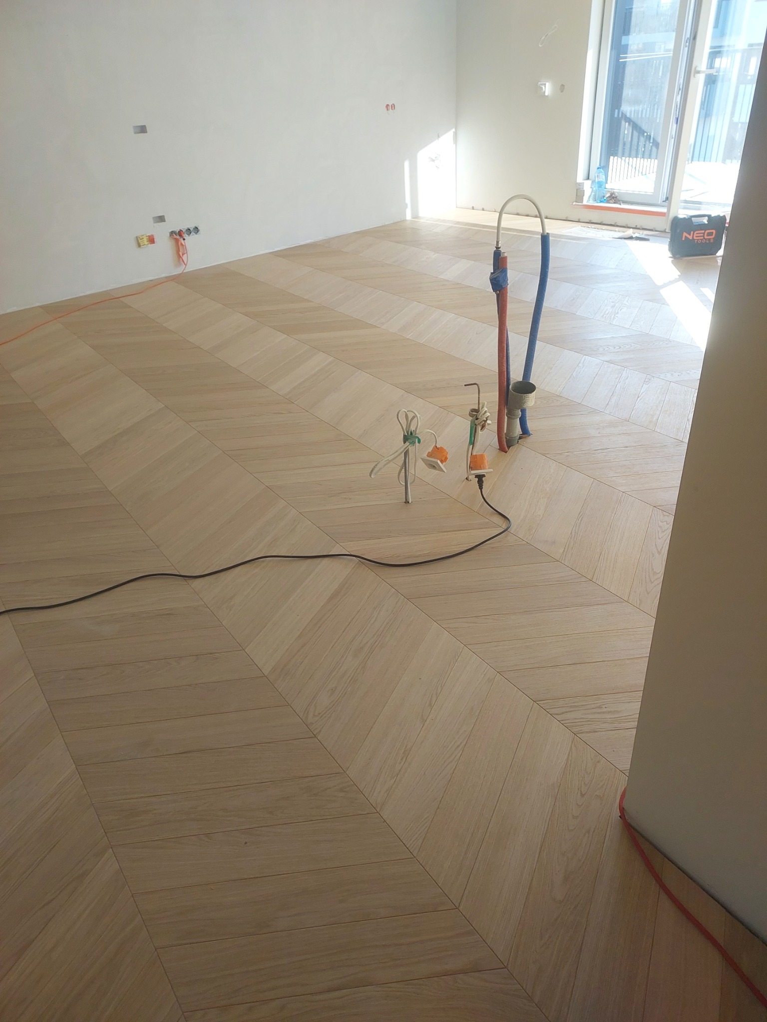 Jasny parkiet ułożony we wzór jodełki w trakcie prac wykończeniowych, widoczne narzędzia i kable na podłodze, fragment okna w tle.