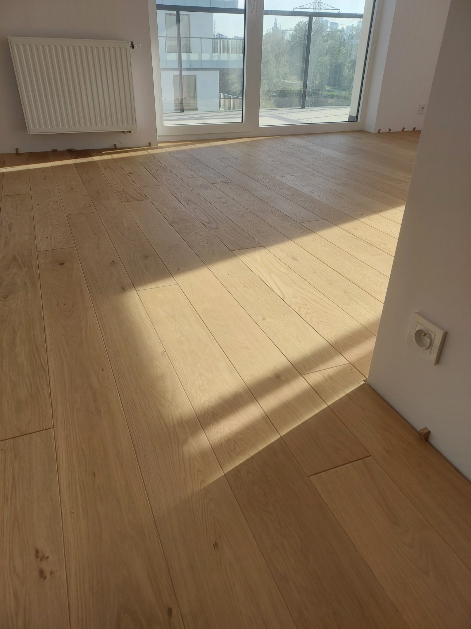 Jasne, drewniane deski parkietowe ułożone w nowym mieszkaniu, widoczne listwy dystansowe przy ścianach i okno balkonowe z widokiem na zieleń.