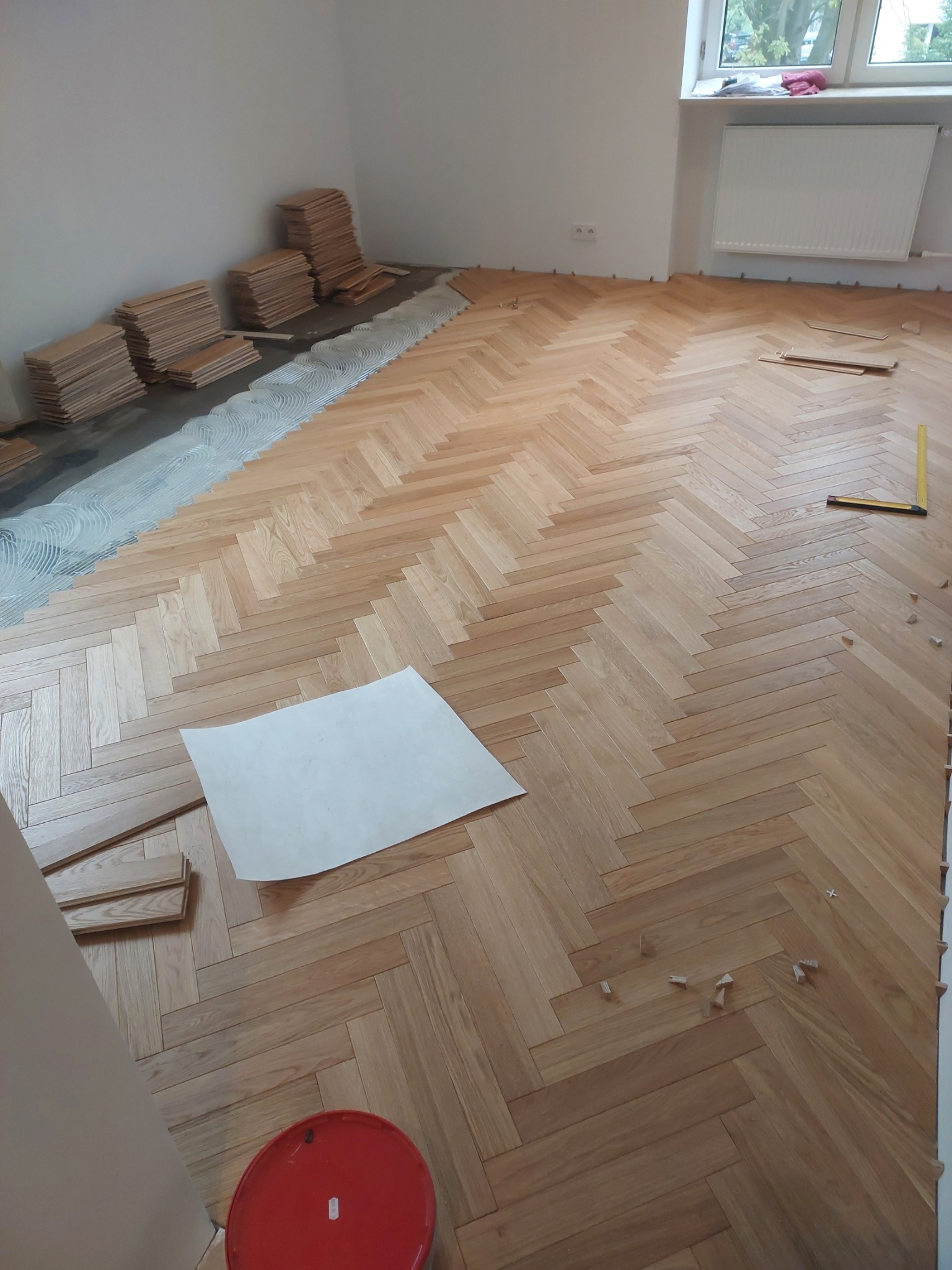 Proces układania parkietu w jodełkę: widok na częściowo ułożoną podłogę, klej, stosy desek, arkusz papieru i narzędzia pomiarowe.