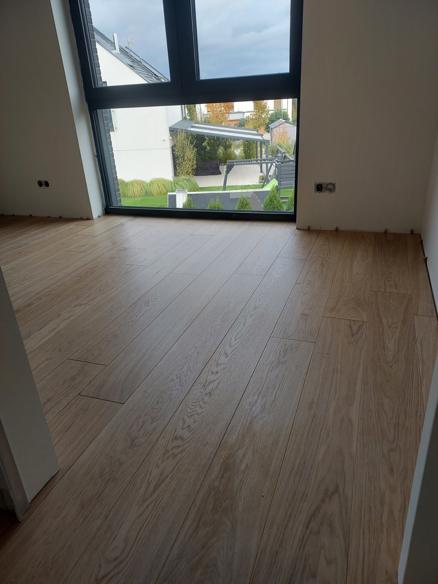 Jasne panele podłogowe ułożone w pokoju z dużym oknem, przez które widać zielony trawnik i nowoczesną architekturę.