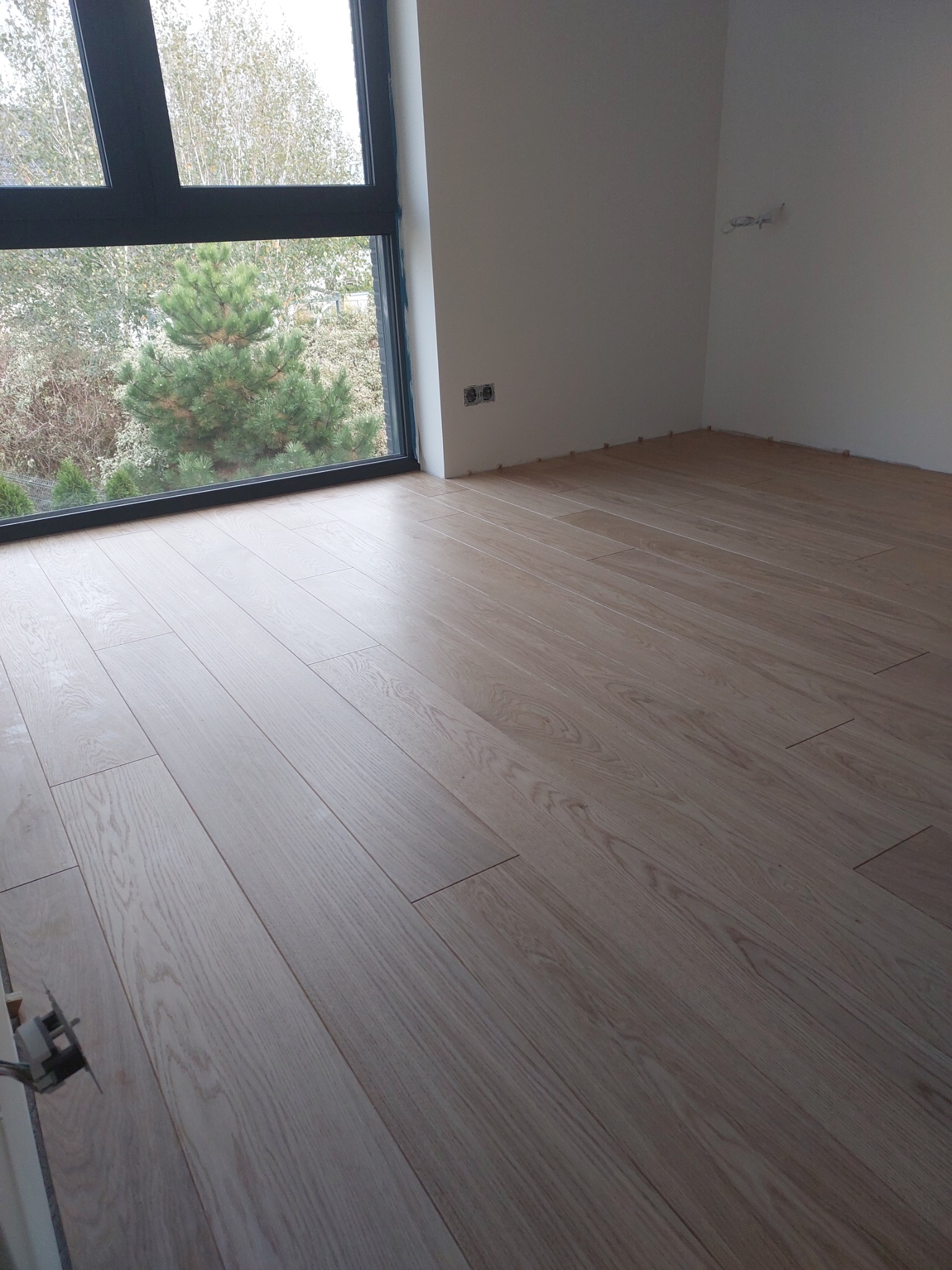 Jasne, drewniane deski podłogowe ułożone w nowym wnętrzu z widokiem na zieleń przez duże okno.