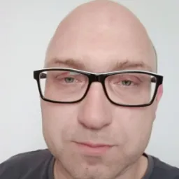 Portret mężczyzny w okularach z czarnymi oprawkami i białymi akcentami na tle jasnej ściany.