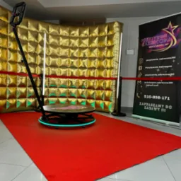 Automatyczna platforma obrotowa 360 stopni z oświetleniem LED na czerwonym dywanie, tło z błyszczących, złotych paneli, obok baner reklamowy.