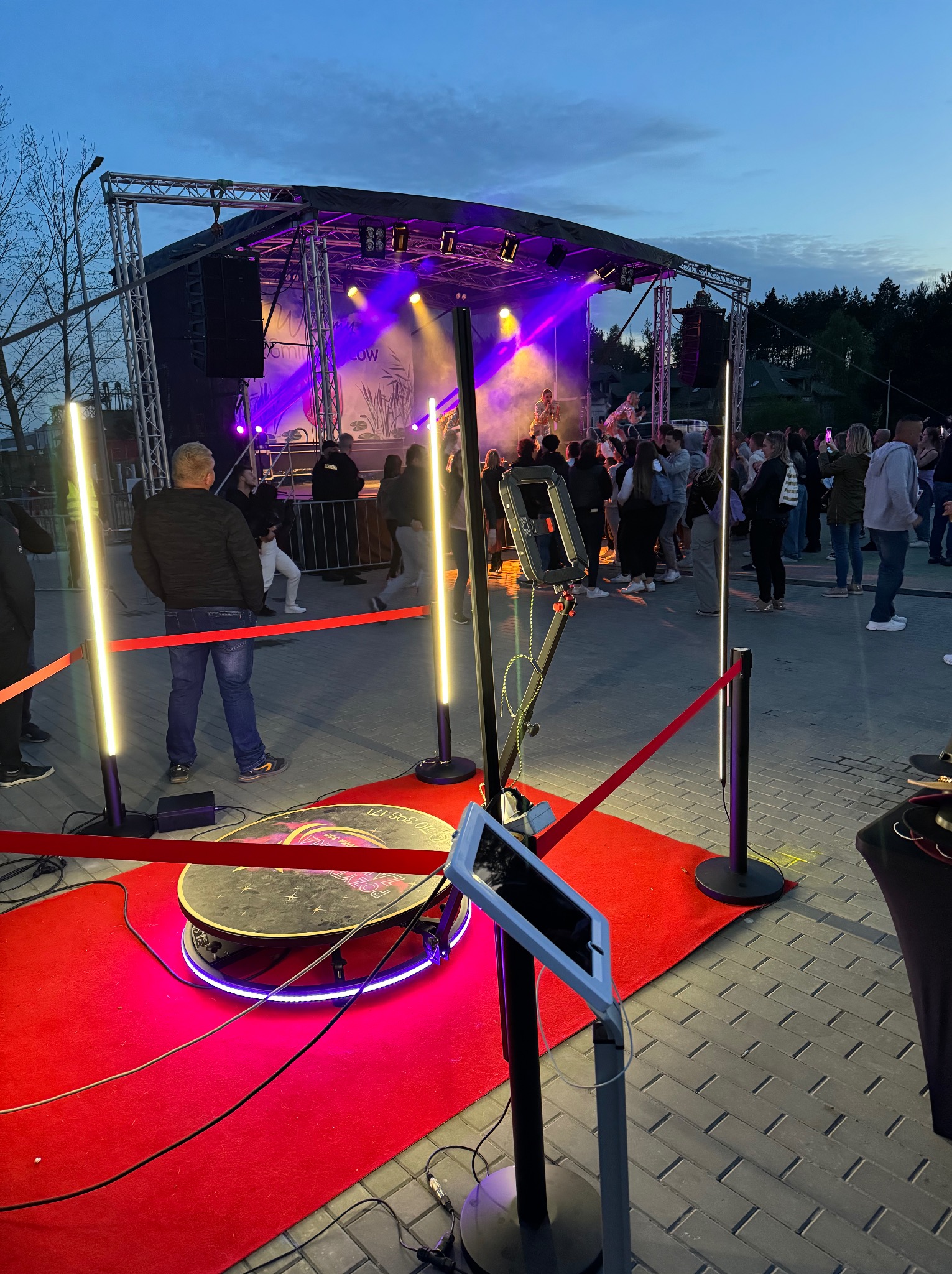 Fotobudka 360 na czerwonym dywanie, otoczona barierkami, w tle scena z występem na imprezie plenerowej o zmierzchu.