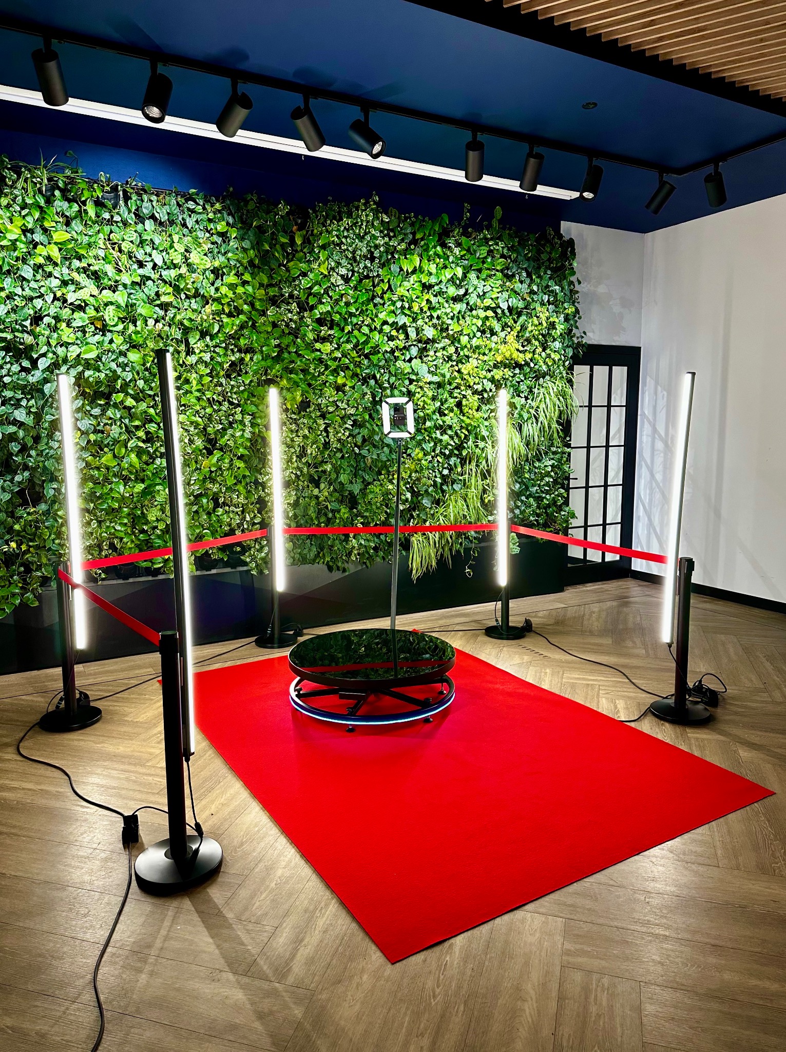 Wnętrze z platformą obrotową do nagrywania filmów 360 stopni, otoczoną czerwonym dywanem i oświetleniem LED, na tle ściany z zielonych roślin.