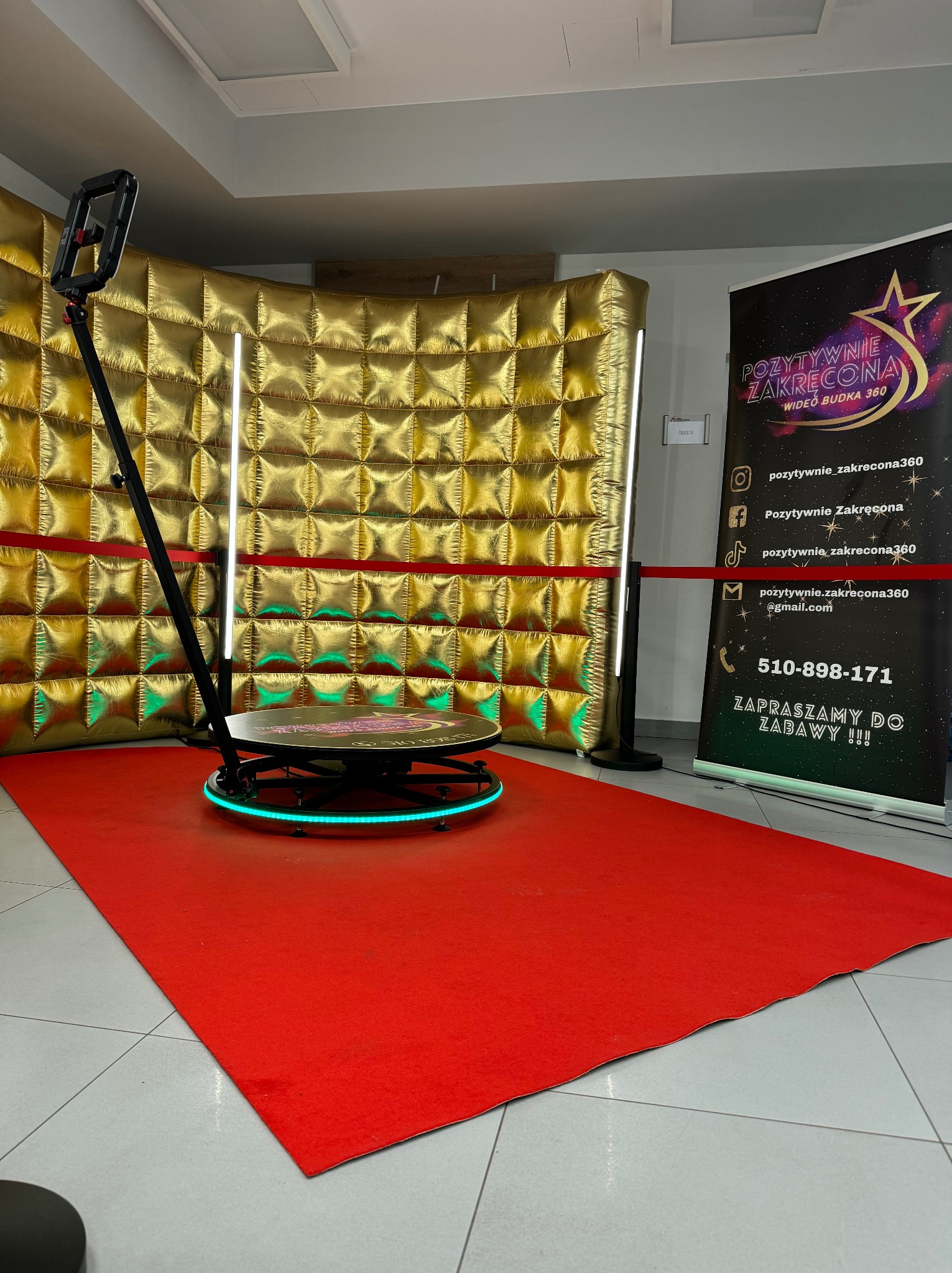 Automatyczna platforma obrotowa 360 stopni z oświetleniem LED na czerwonym dywanie, tło z błyszczących, złotych paneli, obok baner reklamowy.