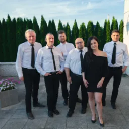 Zespół w czarno-białych strojach pozuje na zewnątrz, z kobietą w czarnej sukience na pierwszym planie, na tle ciemnozielonych krzewów i jasnego nieba.