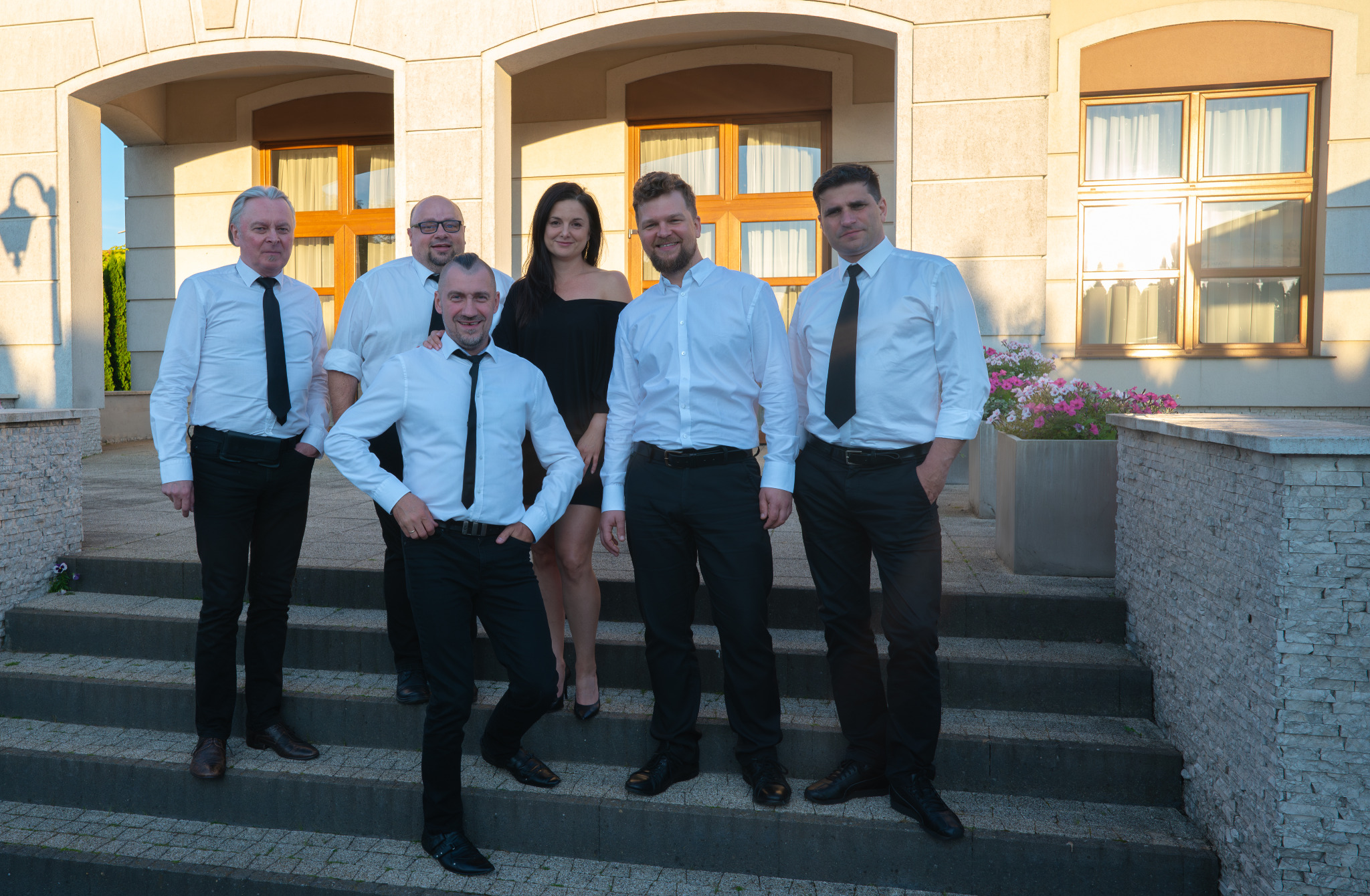 Sześcioosobowa grupa w eleganckich strojach pozuje na schodach przed budynkiem z kwiatami w donicach. Mężczyźni w białych koszulach i czarnych spodniach, z czarnymi krawatami, kobieta w czarnej...