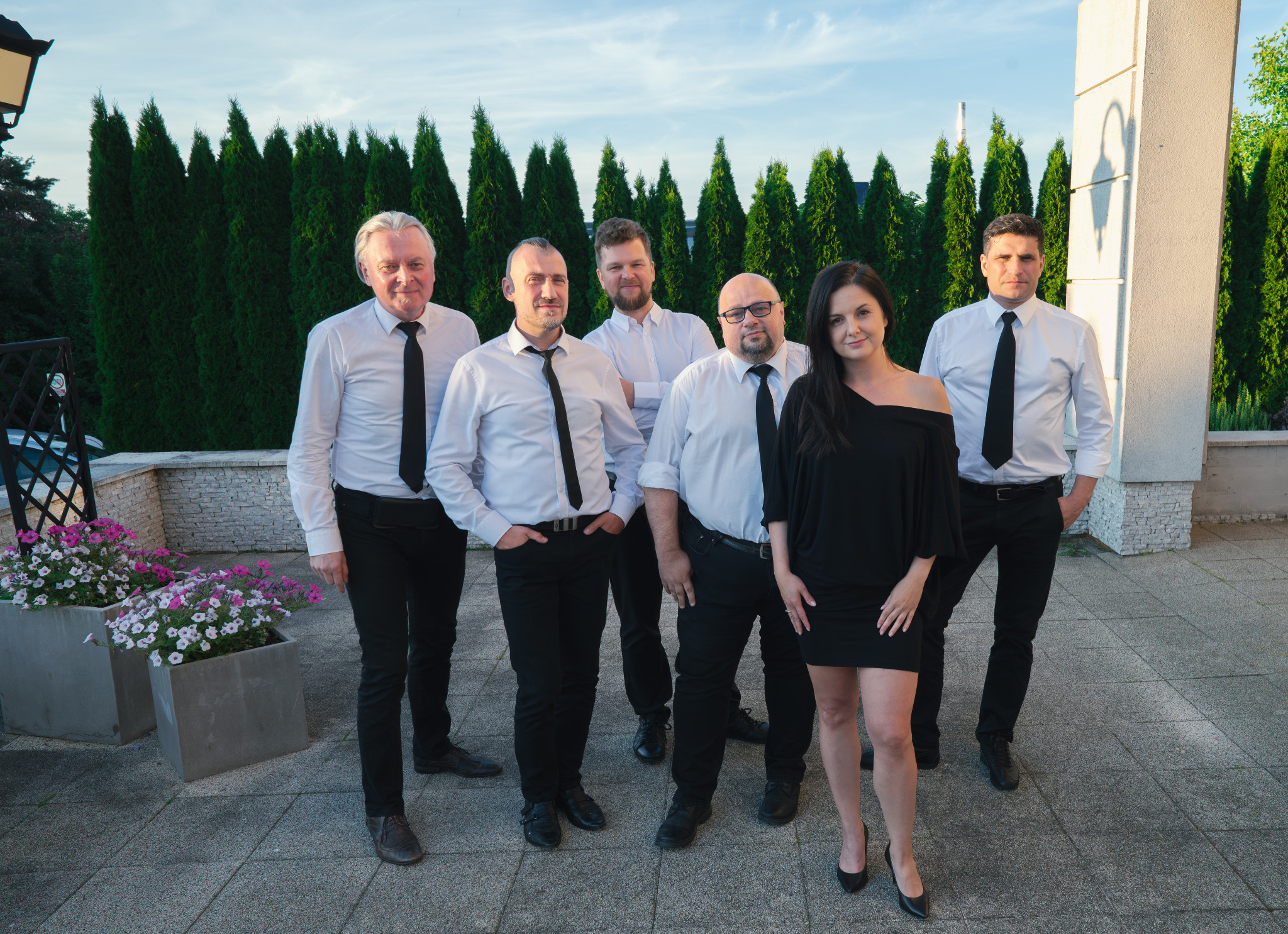 Zespół w czarno-białych strojach pozuje na zewnątrz, z kobietą w czarnej sukience na pierwszym planie, na tle ciemnozielonych krzewów i jasnego nieba.