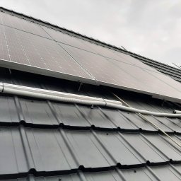 PVE Instalacje Elektryczne Dawid Petrycki - Panele fotowoltaiczne zamontowane na czarnym dachu z dachówki, widoczne szare niebo w tle i biała rura osłonowa na kable.