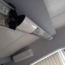 PVE Instalacje Elektryczne Dawid Petrycki - Otwarta obudowa lampy LED na suficie z widocznymi elementami elektrycznymi i złączkami, obok klimatyzator i żaluzje.