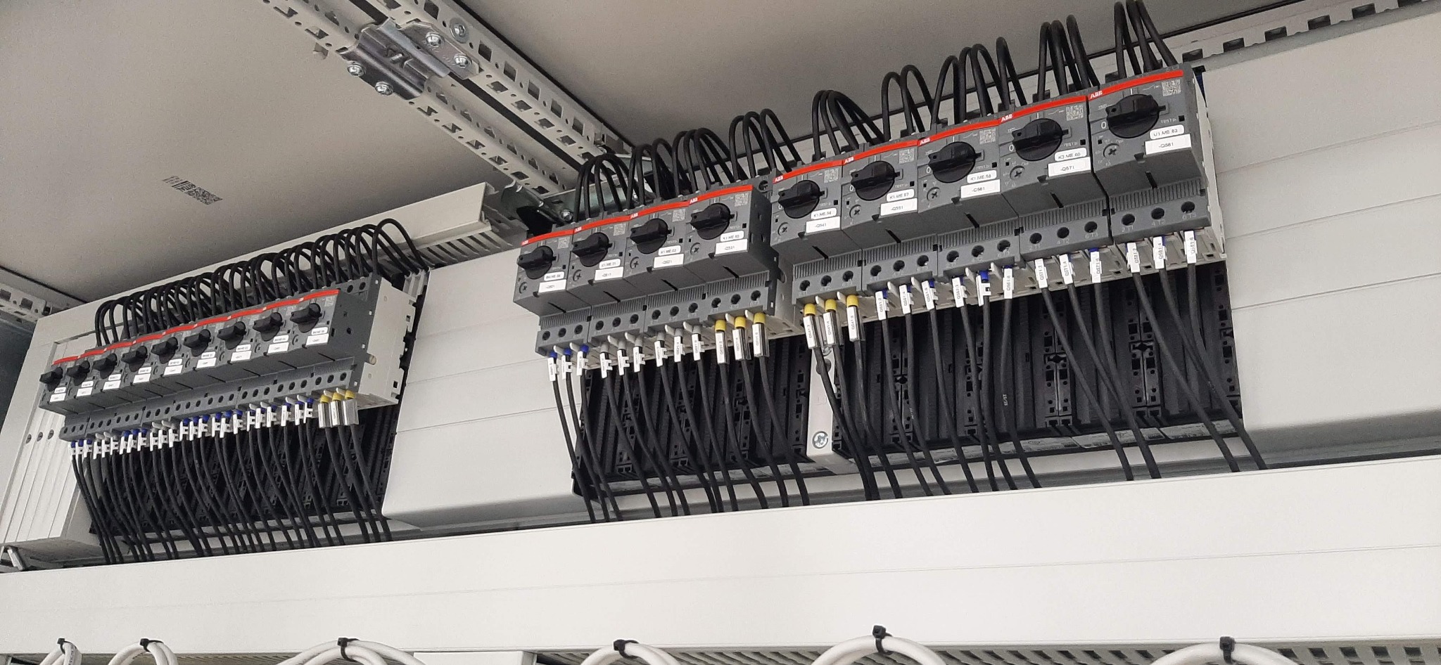 Rozbudowana rozdzielnica elektryczna z widocznymi bezpiecznikami, wyłącznikami i licznymi przewodami podłączonymi do listew zaciskowych, umieszczona w białej szafie sterowniczej.