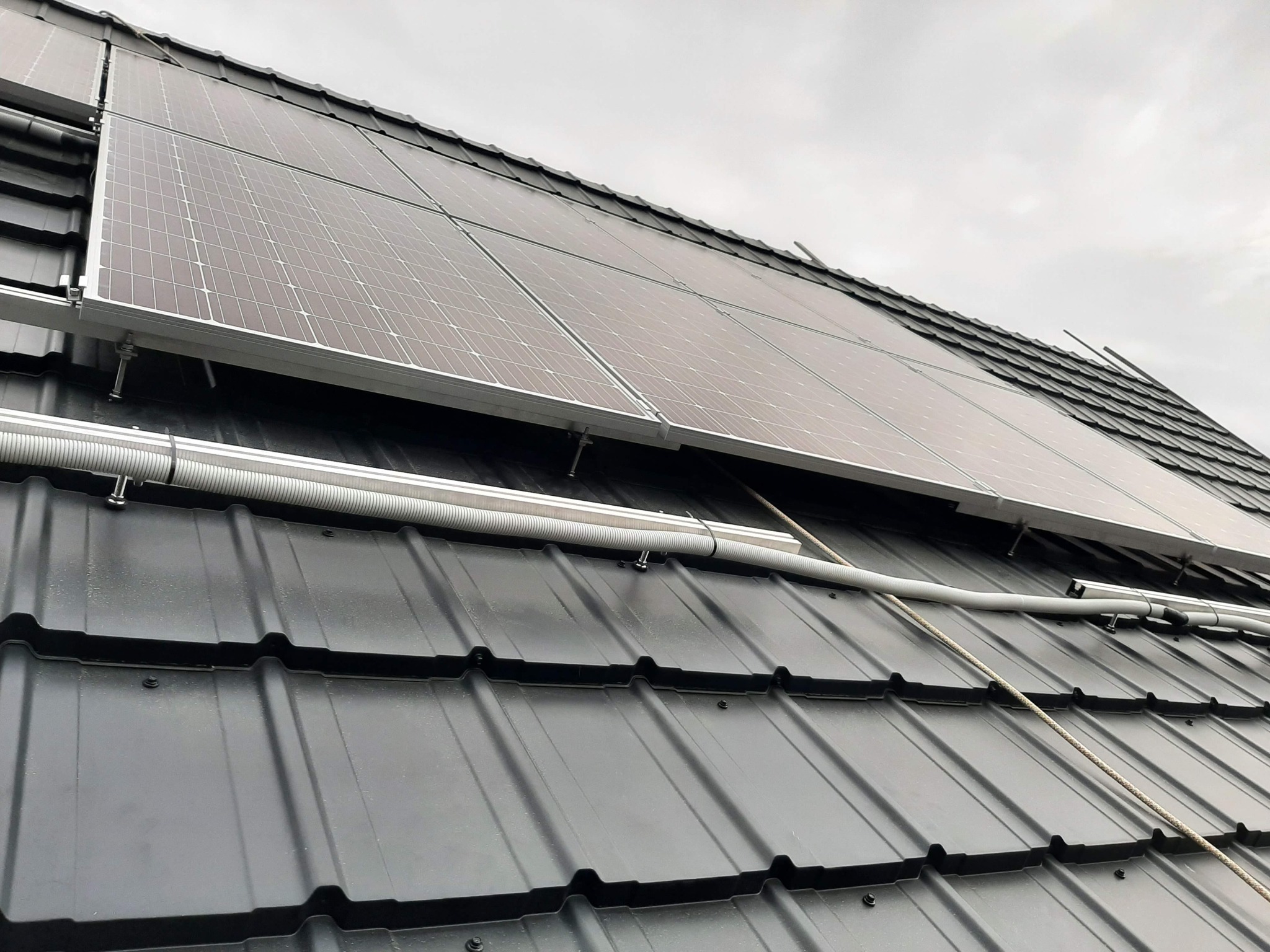 Panele fotowoltaiczne zamontowane na czarnym dachu z dachówki, widoczne szare niebo w tle i biała rura osłonowa na kable.