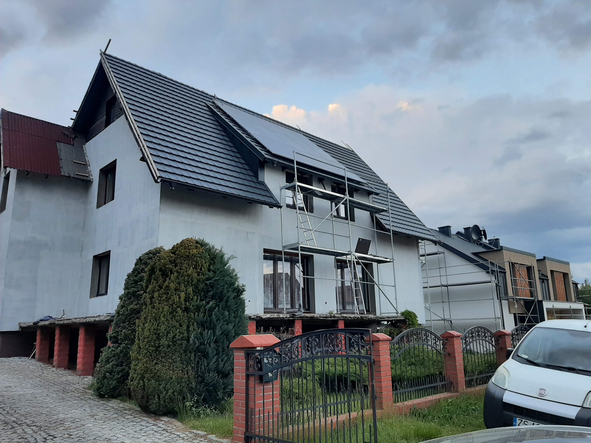 Dom jednorodzinny z białym tynkiem i szarą dachówką, na dachu widoczne panele słoneczne, przy ścianie rusztowanie, a w tle pochmurne niebo.